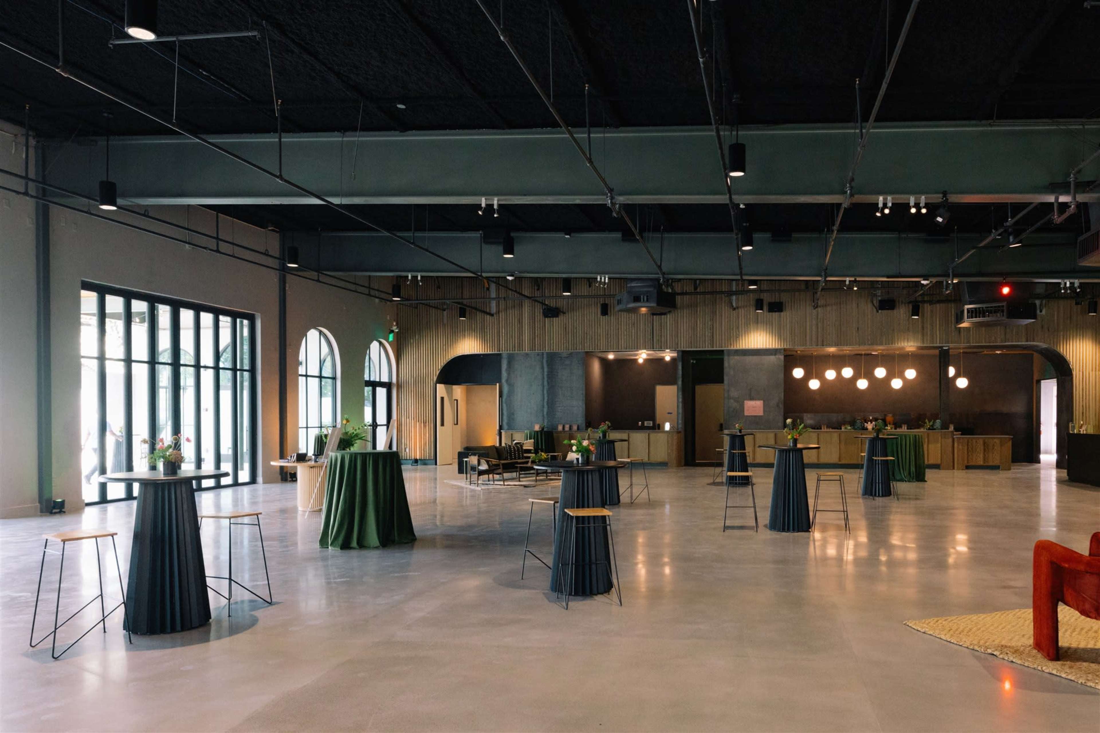 The image shows a spacious, modern event venue featuring high ceilings, circular tables with black and green skirts, and a subdued color palette with minimalist decor.