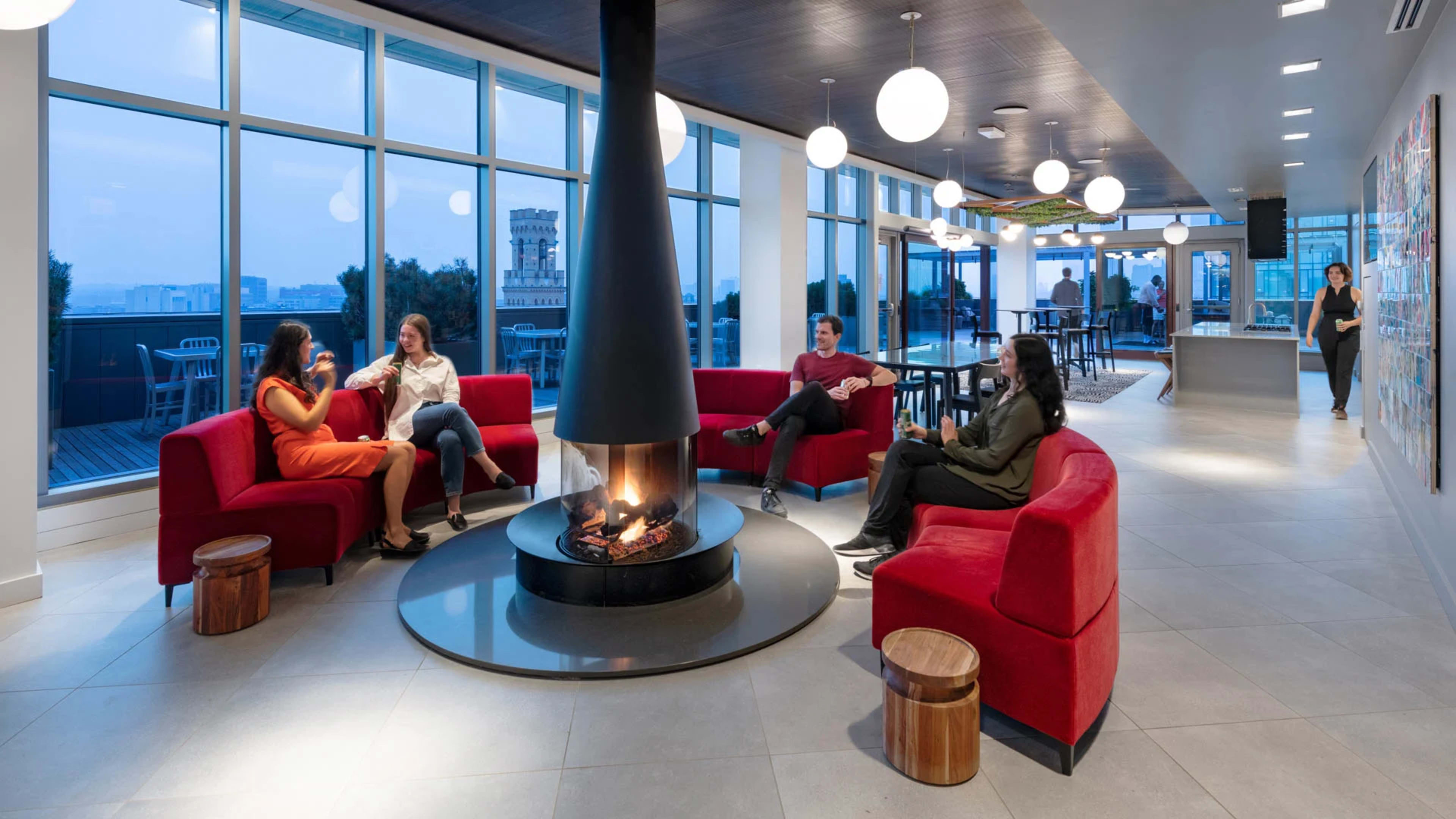 A modern lounge area with comfortable seating around a central fireplace, large windows providing city views, and a few people engaged in conversation.