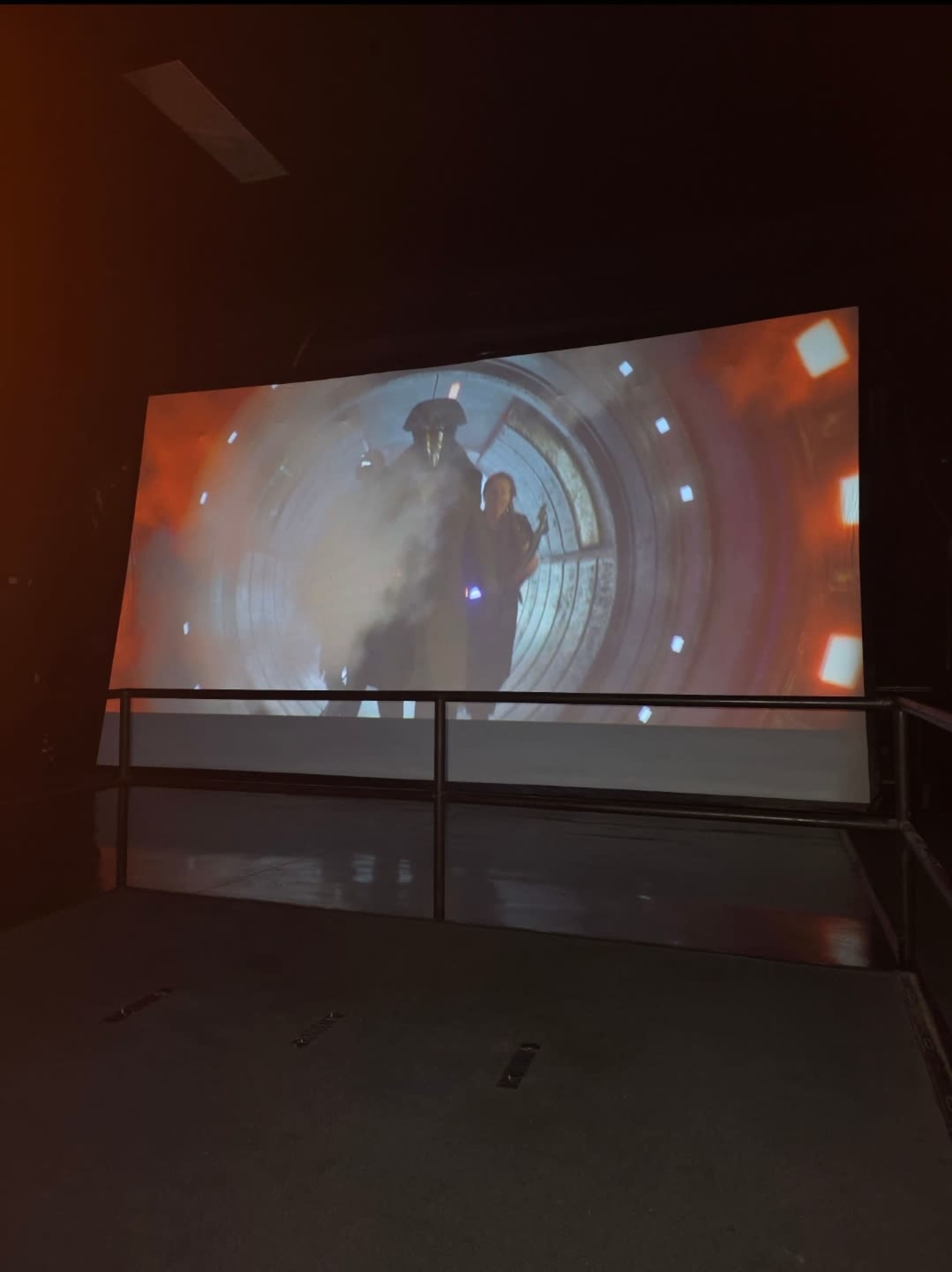 A large curved screen displays a scene from a sci-fi movie featuring two characters emerging from a portal surrounded by smoke and lights.