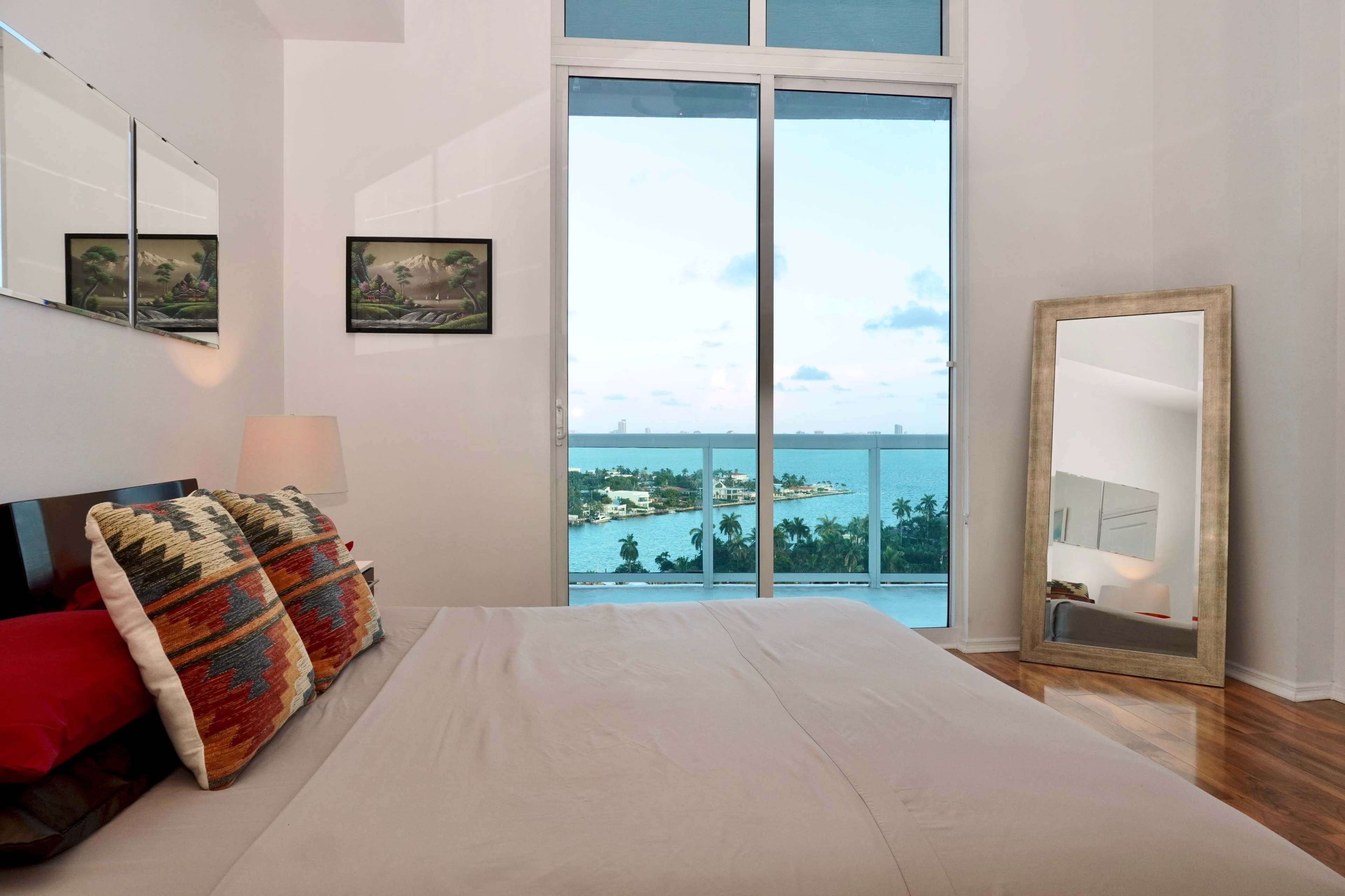 A bedroom features a large window overlooking a body of water, with two paintings on the wall and a mirror beside the bed.