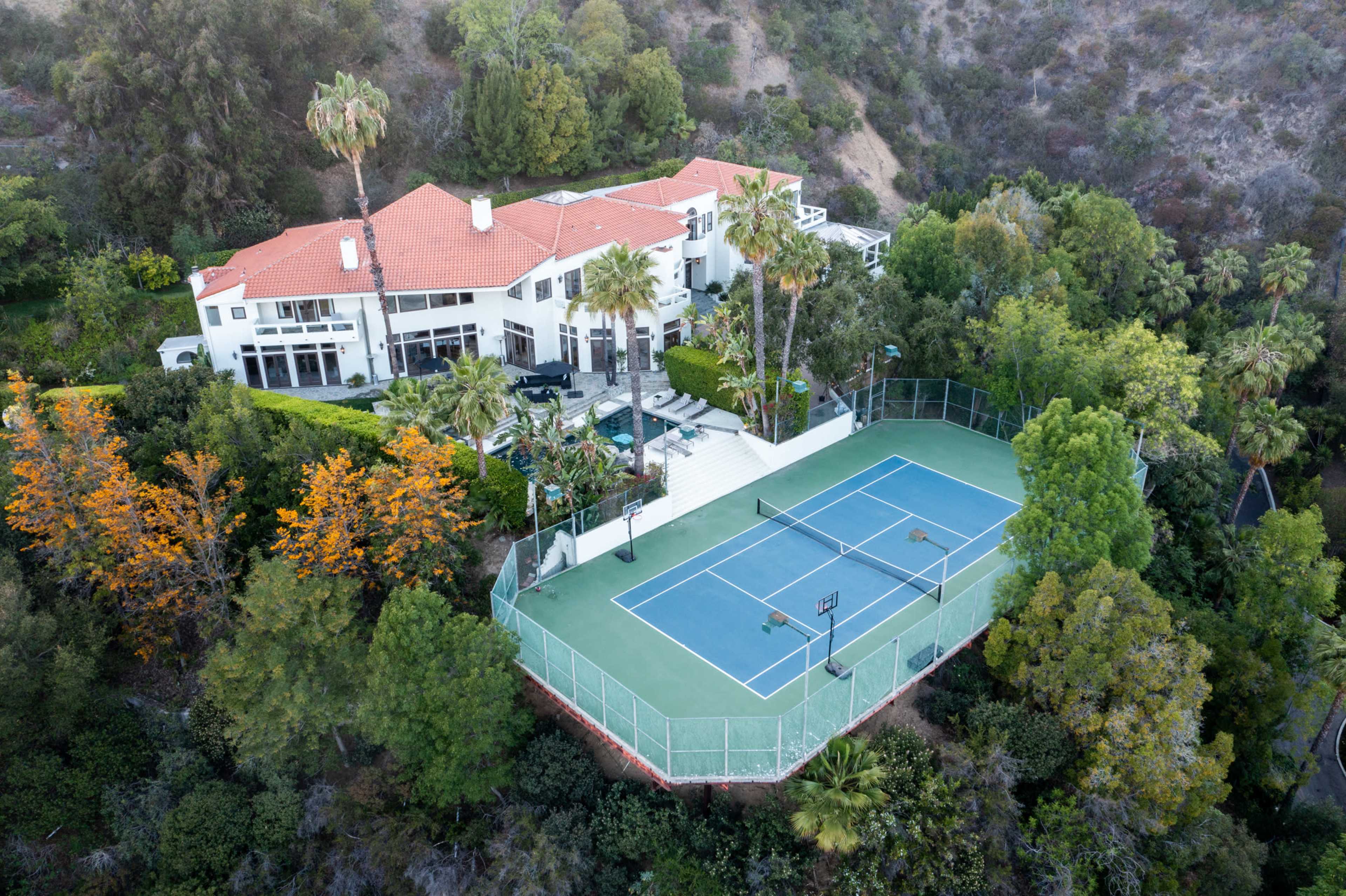 A large house with a red-tiled roof surrounded by greenery and a tennis court nearby.