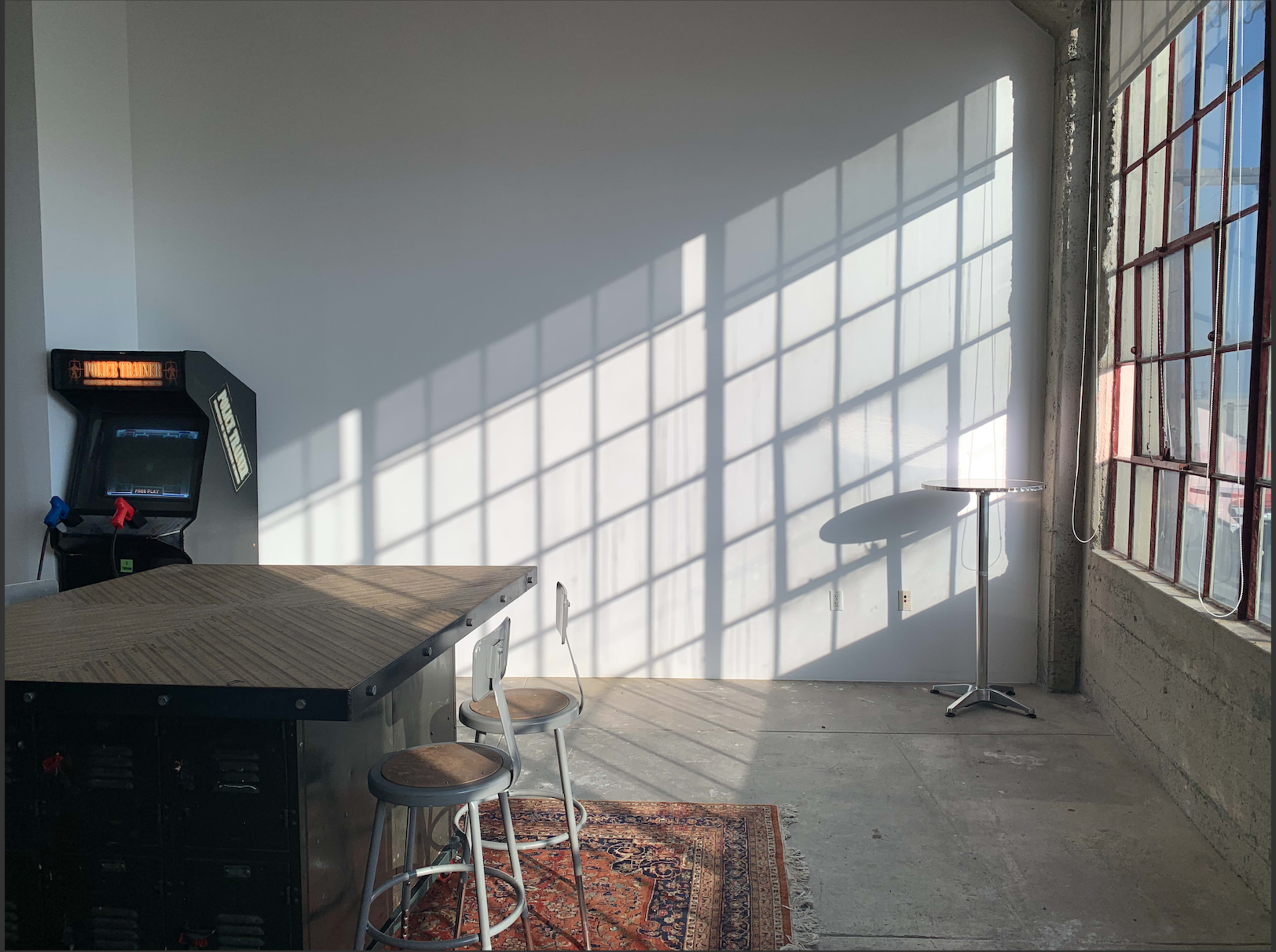 A sunlit room with a wooden bar, two stools, an arcade machine in the corner, and a small table near a large window casting shadows on the wall.