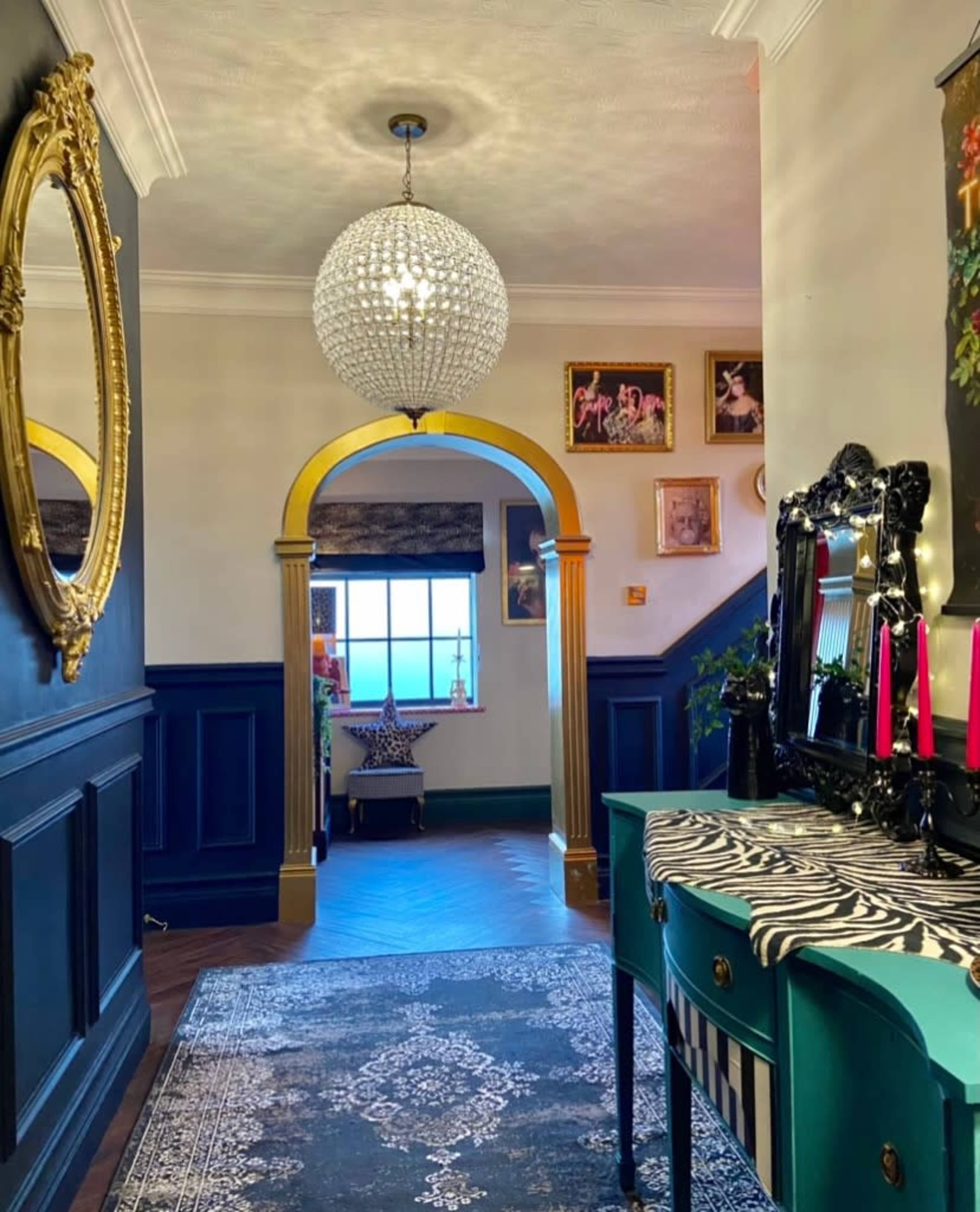 A hallway with dark blue walls, a round chandelier, and an ornate mirror, leading to an archway and a staircase.