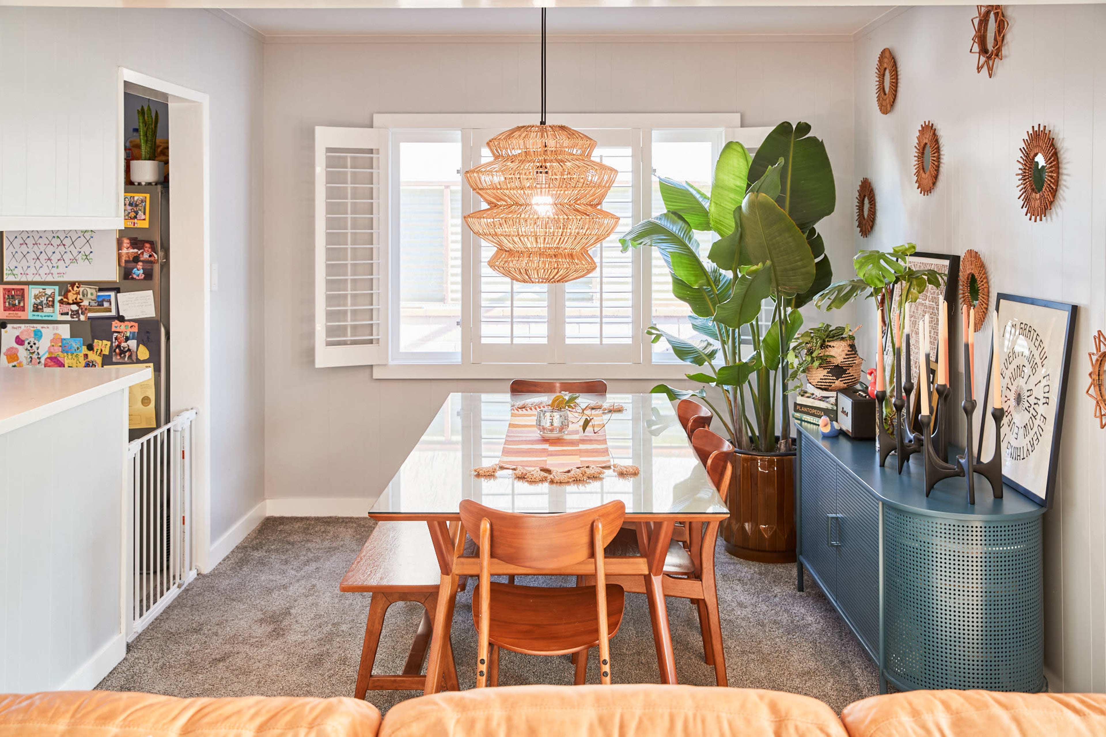 The dining area features a large table surrounded by wooden chairs, a woven pendant light above, and decorative plants and wall art.