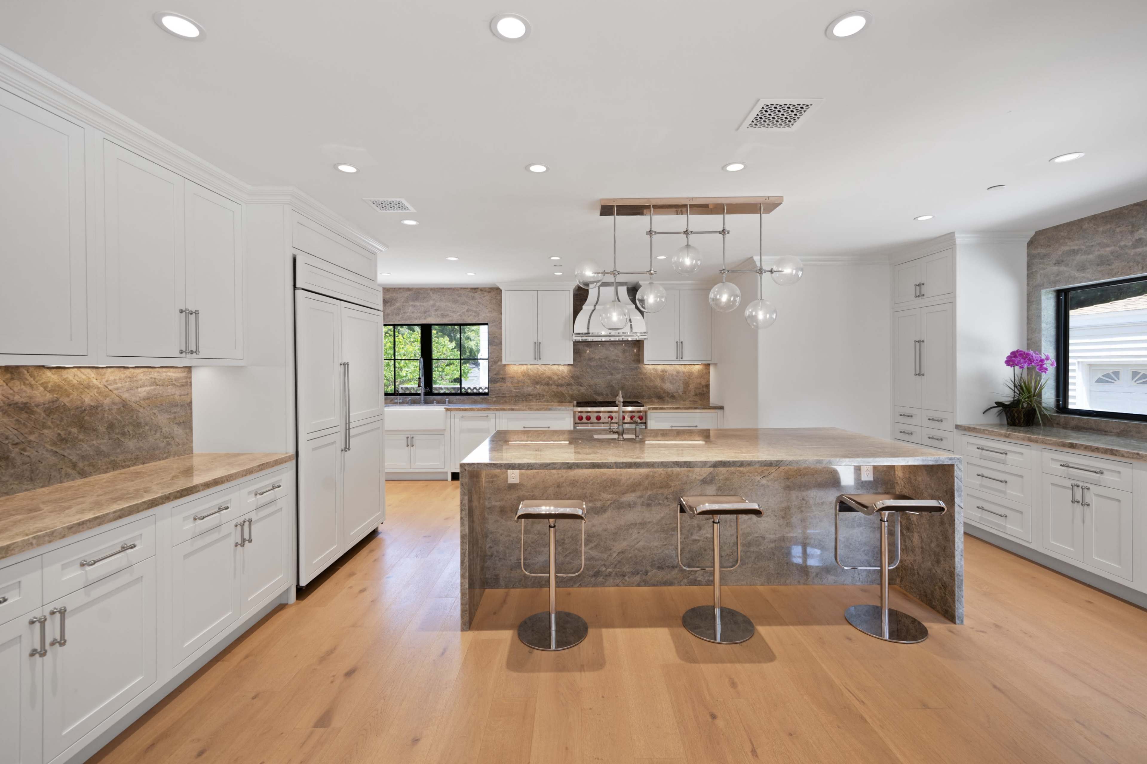 A modern kitchen features white cabinetry, a large marble island with bar stools, and stainless steel appliances under bright overhead lighting.