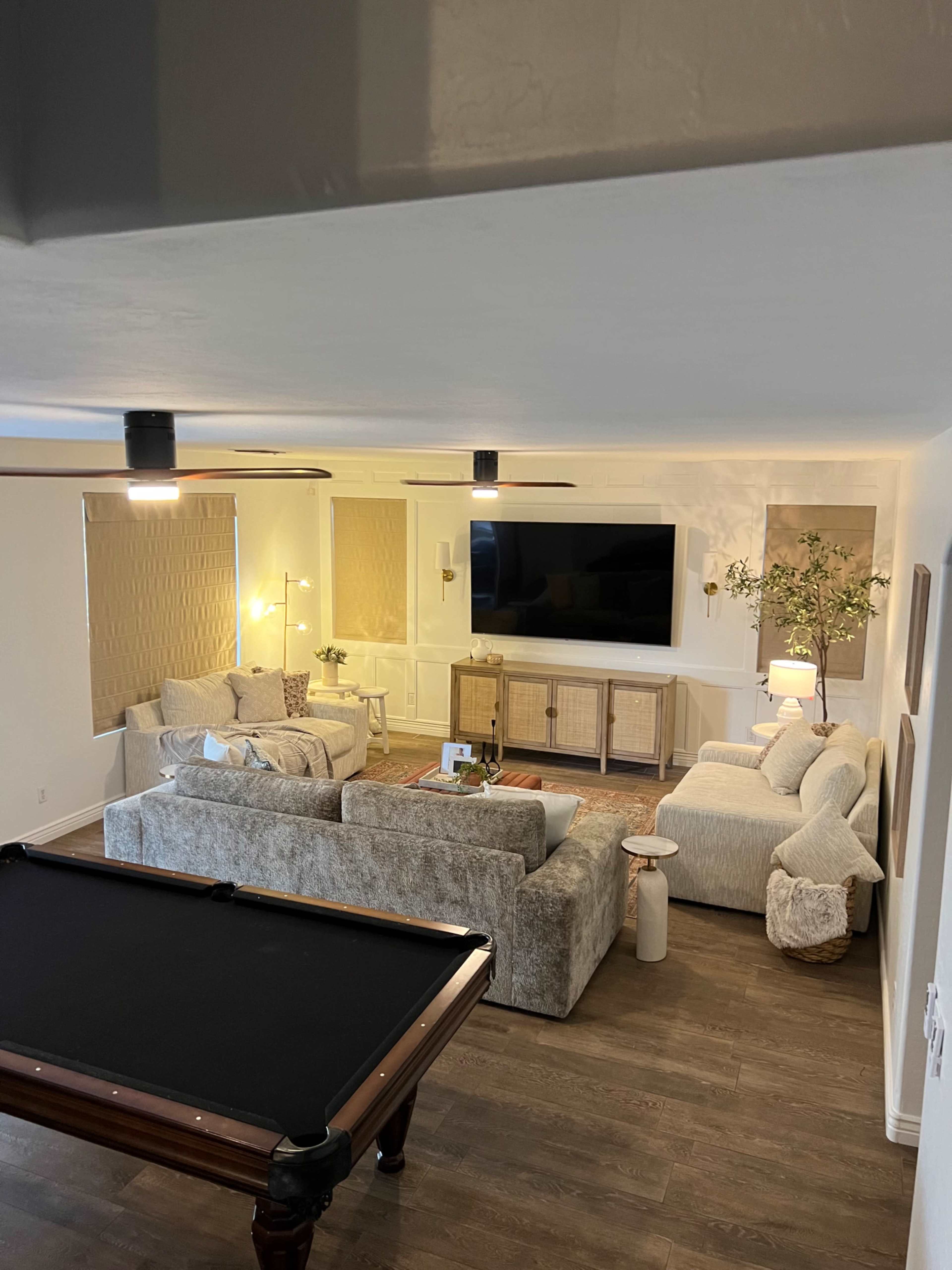 The image shows a modern living room featuring a sectional sofa, a coffee table, a large television mounted on the wall, and a pool table in the foreground.