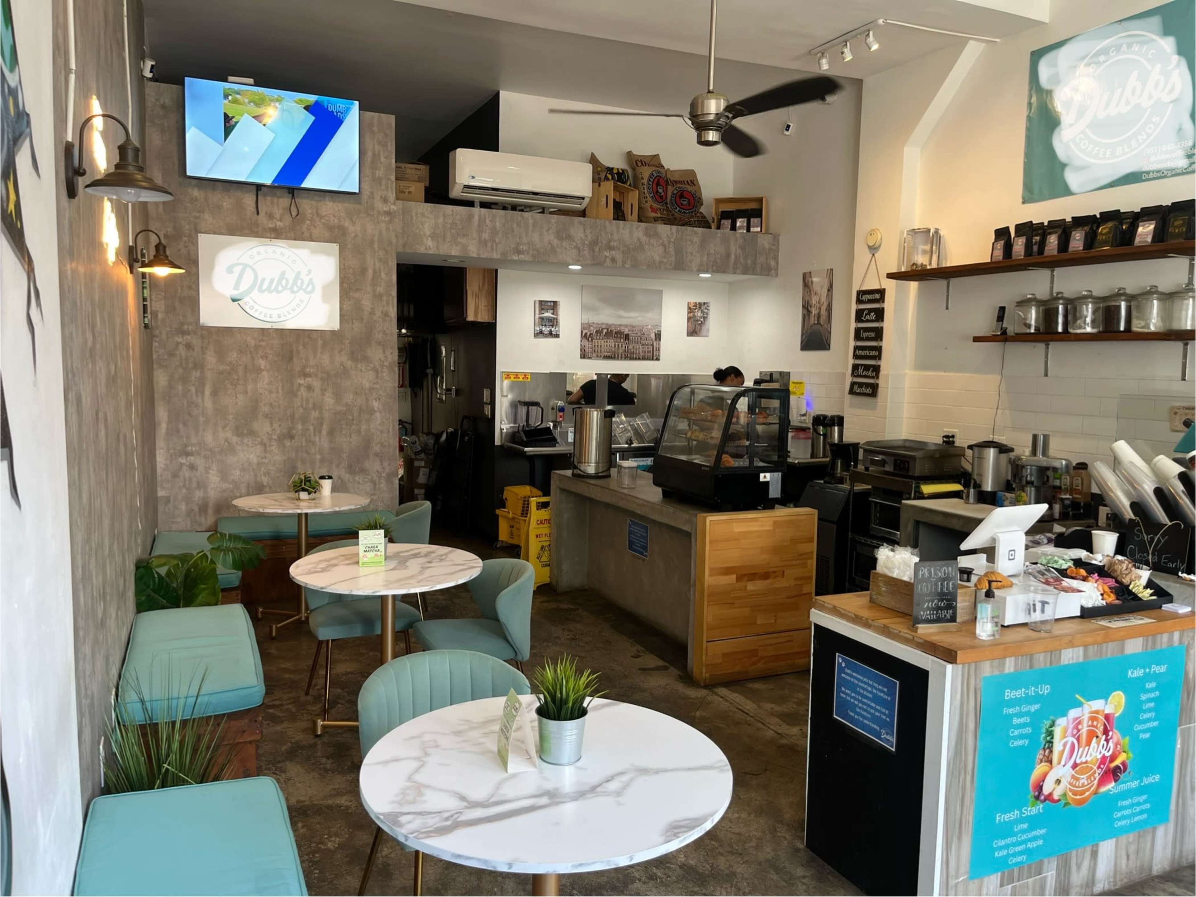 The image shows a cozy cafe interior with marble-topped tables, a small counter displaying coffee equipment, and a wall-mounted television.