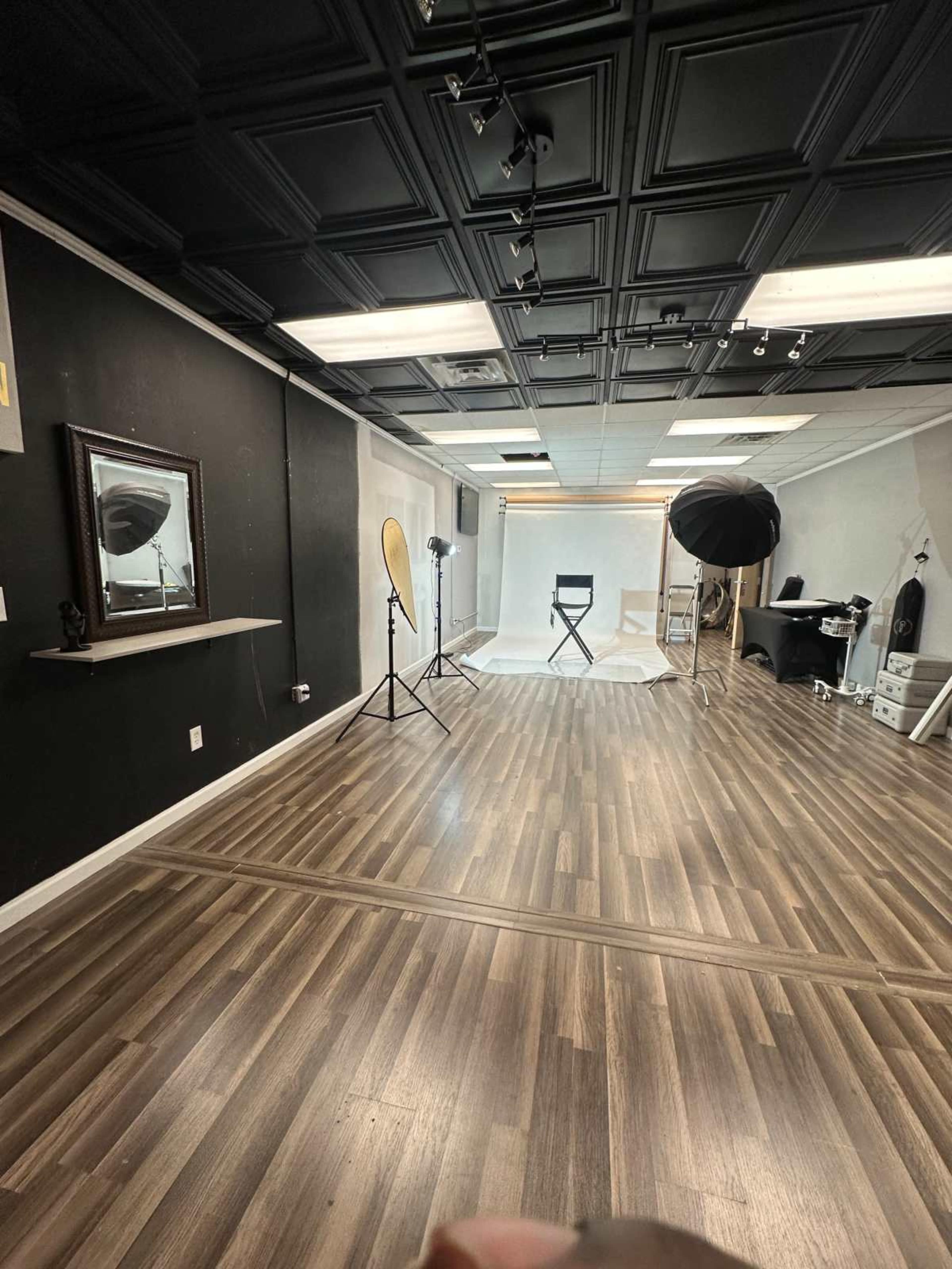 The image shows a spacious photography studio with a light-colored backdrop, lighting equipment, a black chair, and wooden flooring.