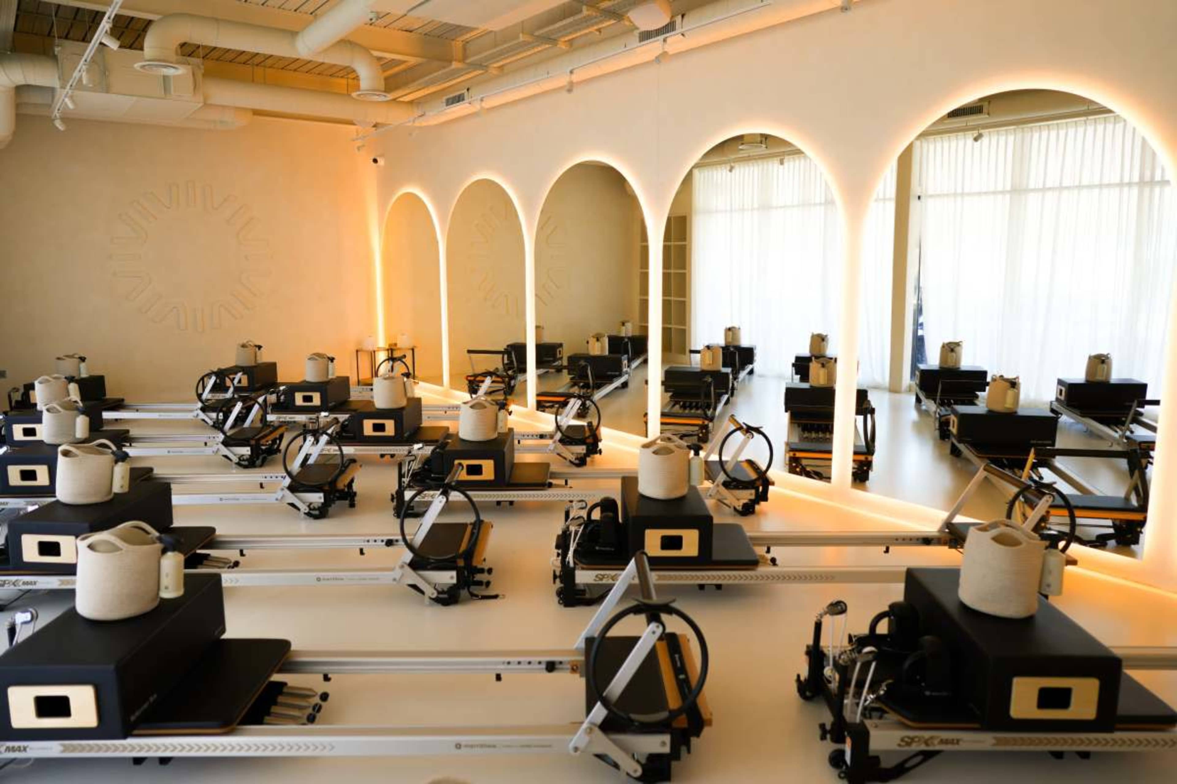 The image shows a modern fitness studio equipped with several reformer Pilates machines arranged in rows against a softly lit backdrop with arched mirrors.