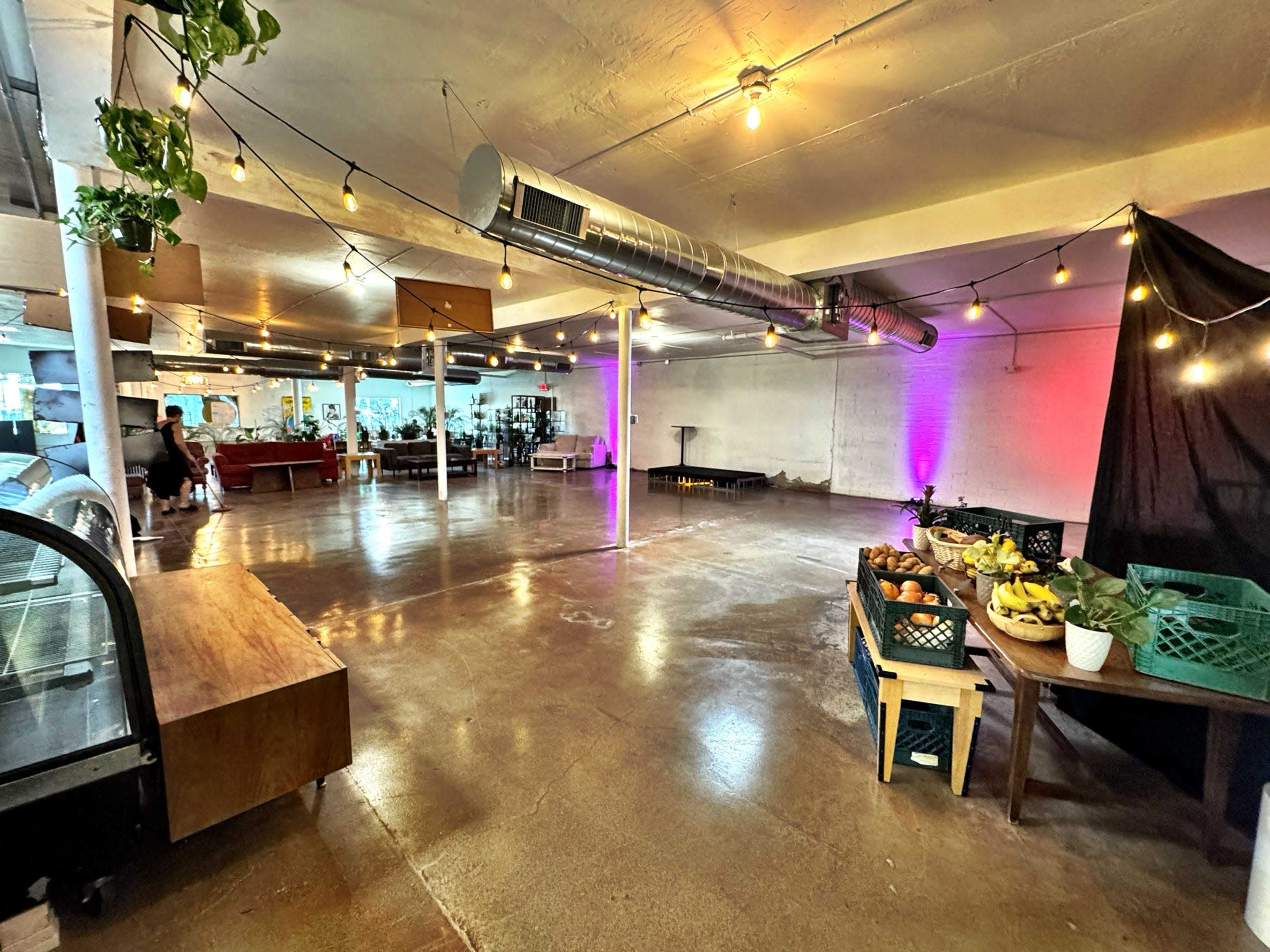 The image shows an empty indoor space with polished concrete floors, string lights hanging from the ceiling, tables with baskets of fruits, and a gathering area in the background.