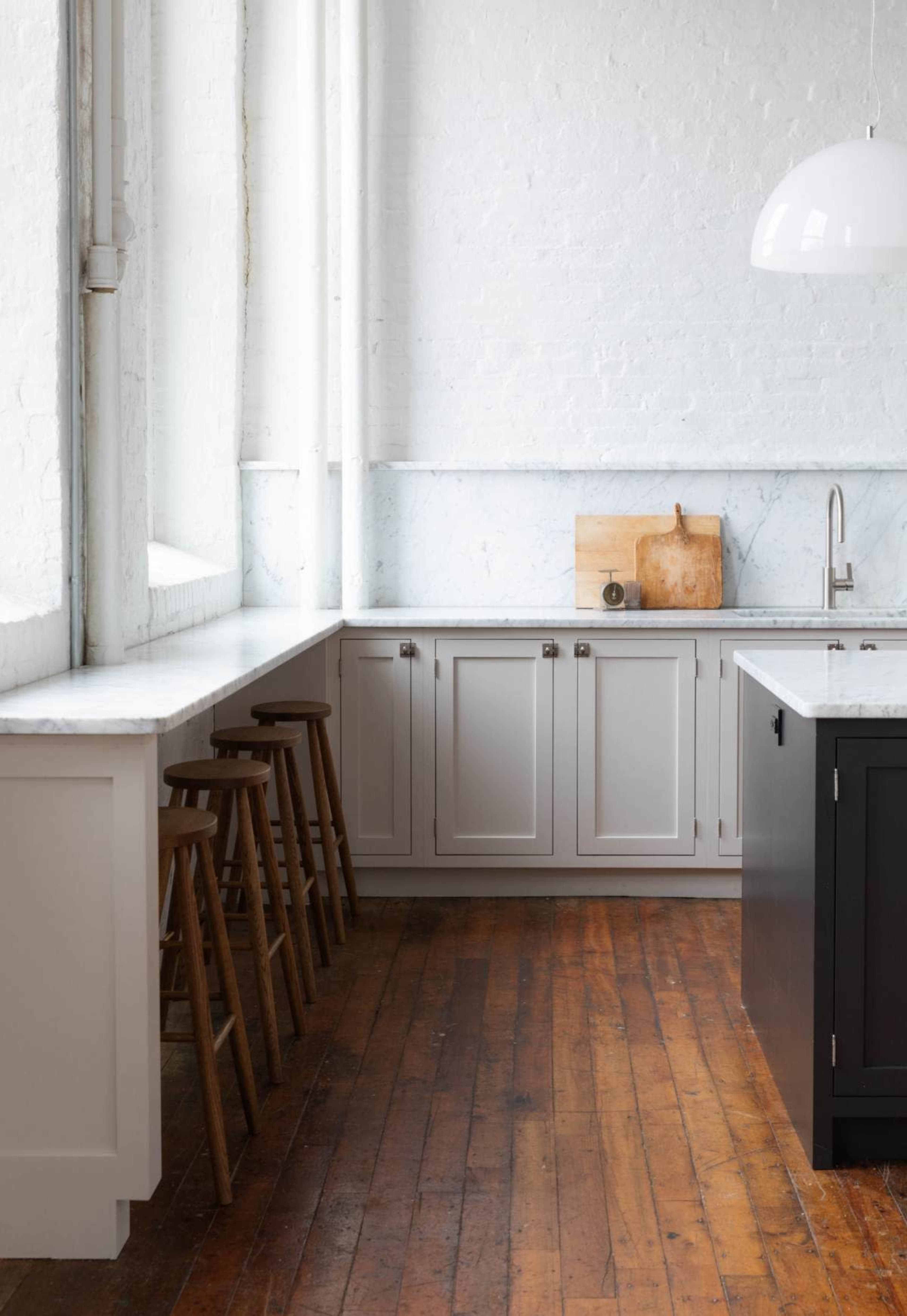 A spacious kitchen features white walls, marble countertops, and wooden flooring, with stools arranged along a long countertop.
