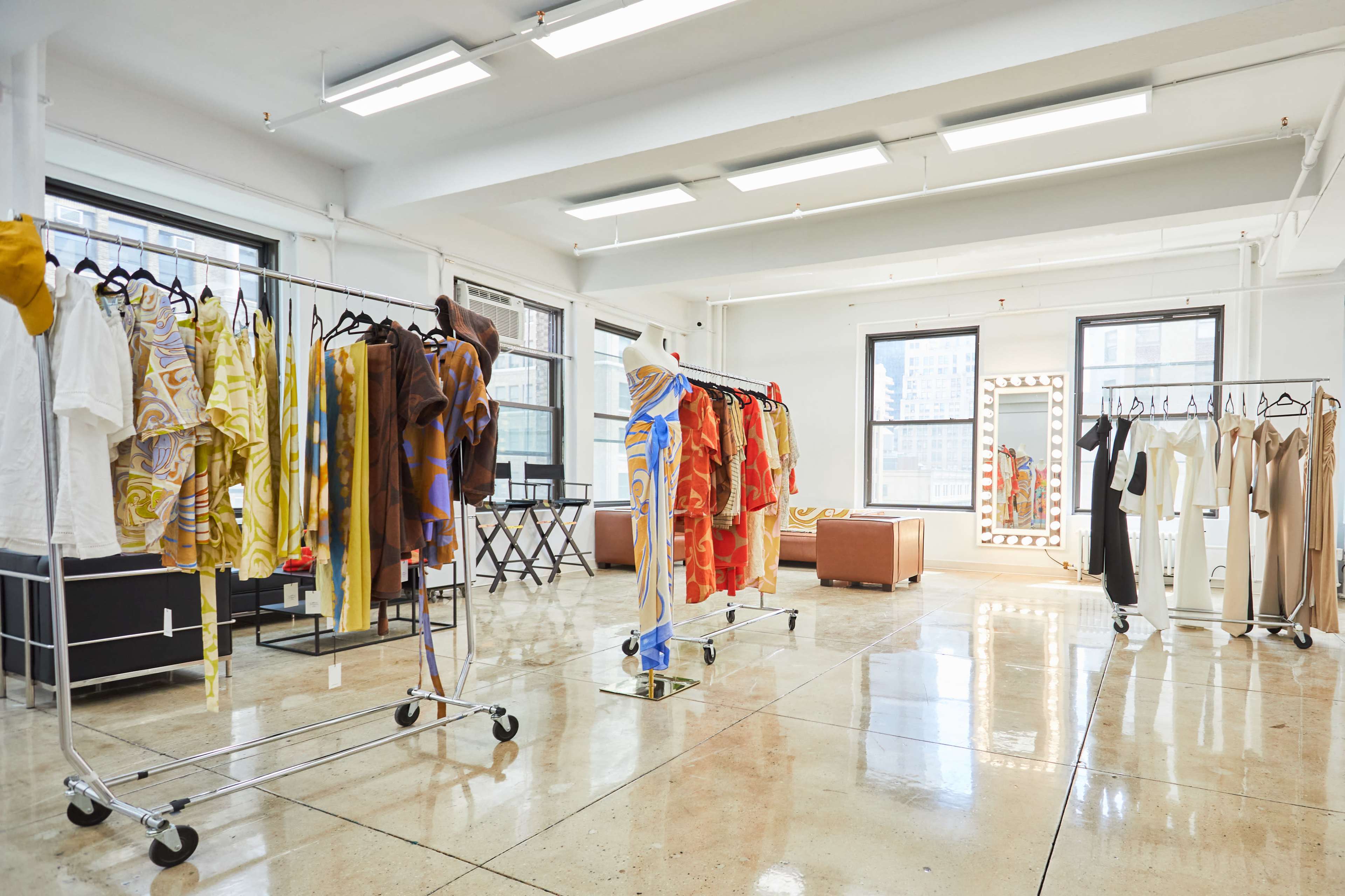 A brightly lit clothing showroom features several racks of colorful garments and a large mirror in the background.
