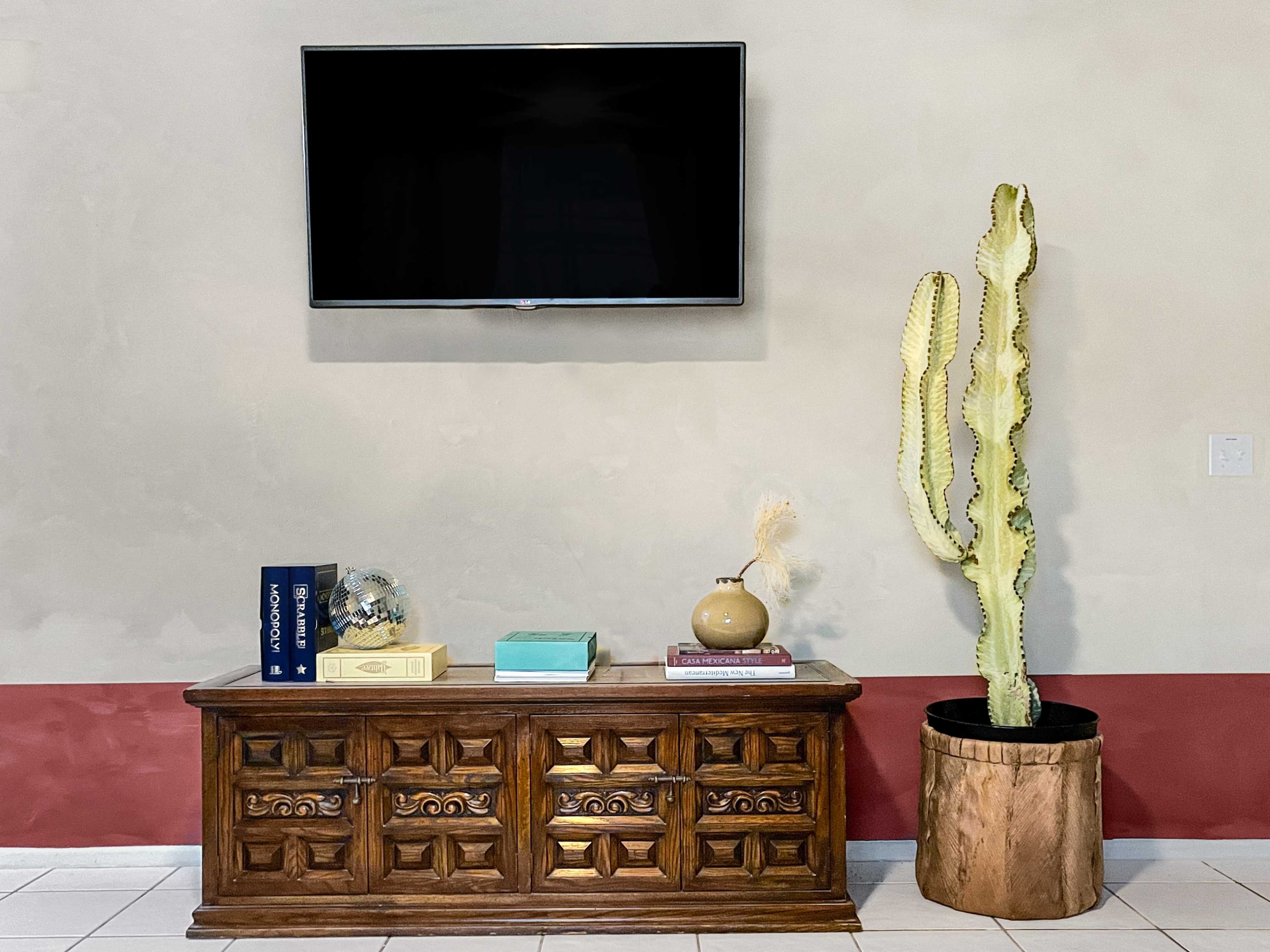 A wooden cabinet with ornate detailing sits beneath a wall-mounted television, accompanied by a tall cactus in a pot and various decorative items.