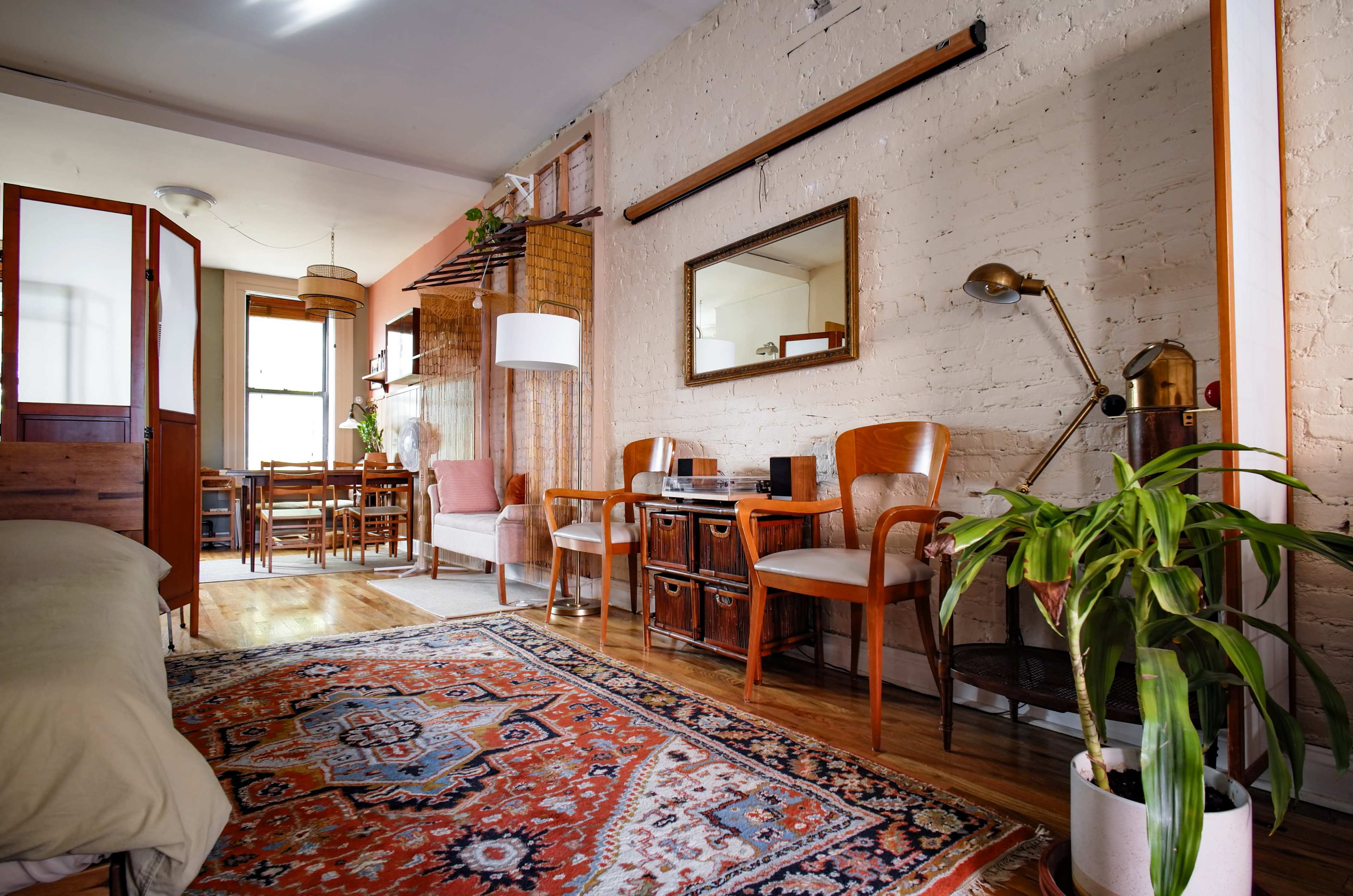 The image shows a cozy interior with a living area featuring wooden furniture, a decorative rug, and plants, illuminated by natural light.