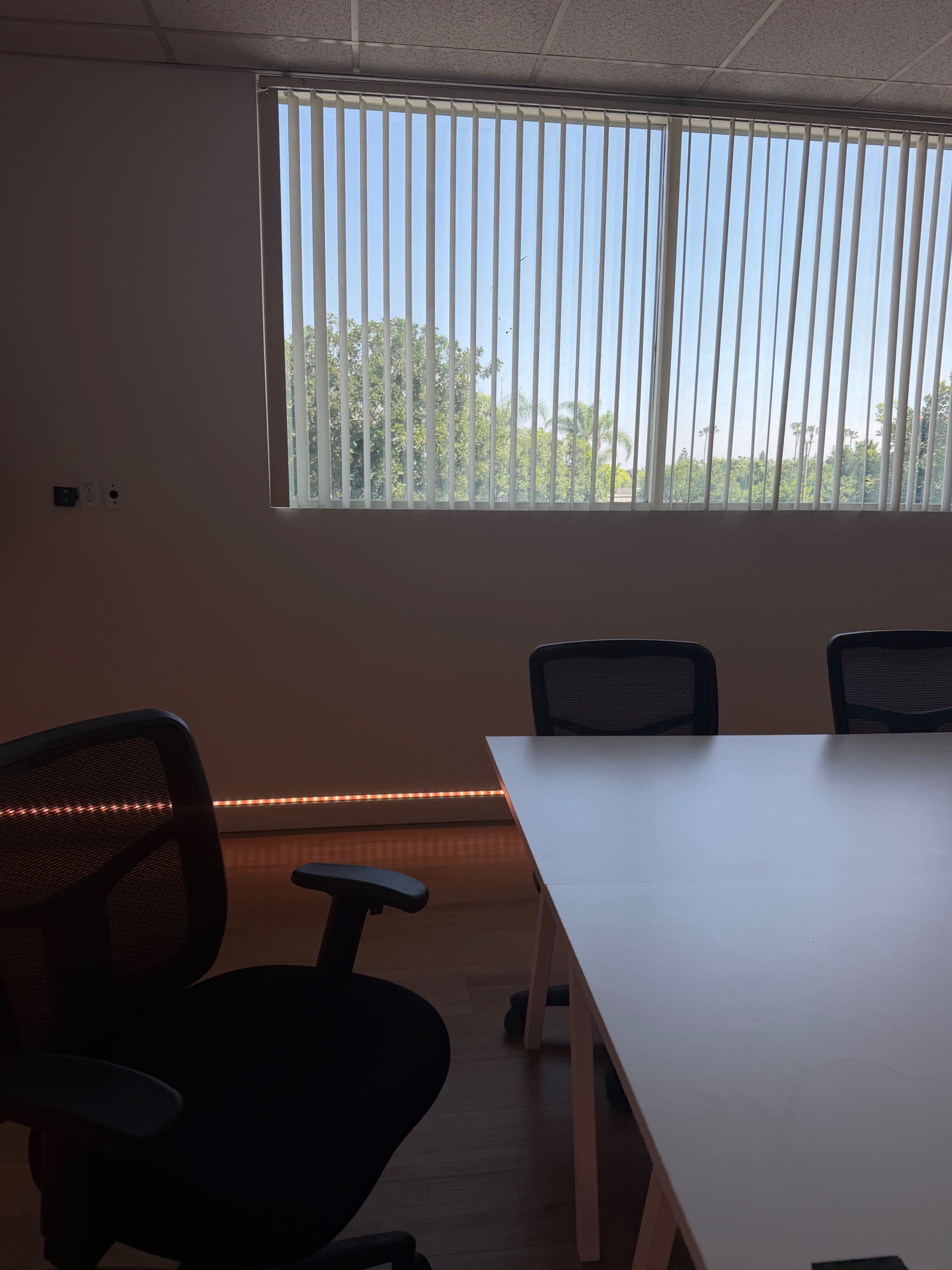 The image shows a modern office interior featuring a white table and black chairs, with large vertical blinds covering a window that reveals greenery outside.