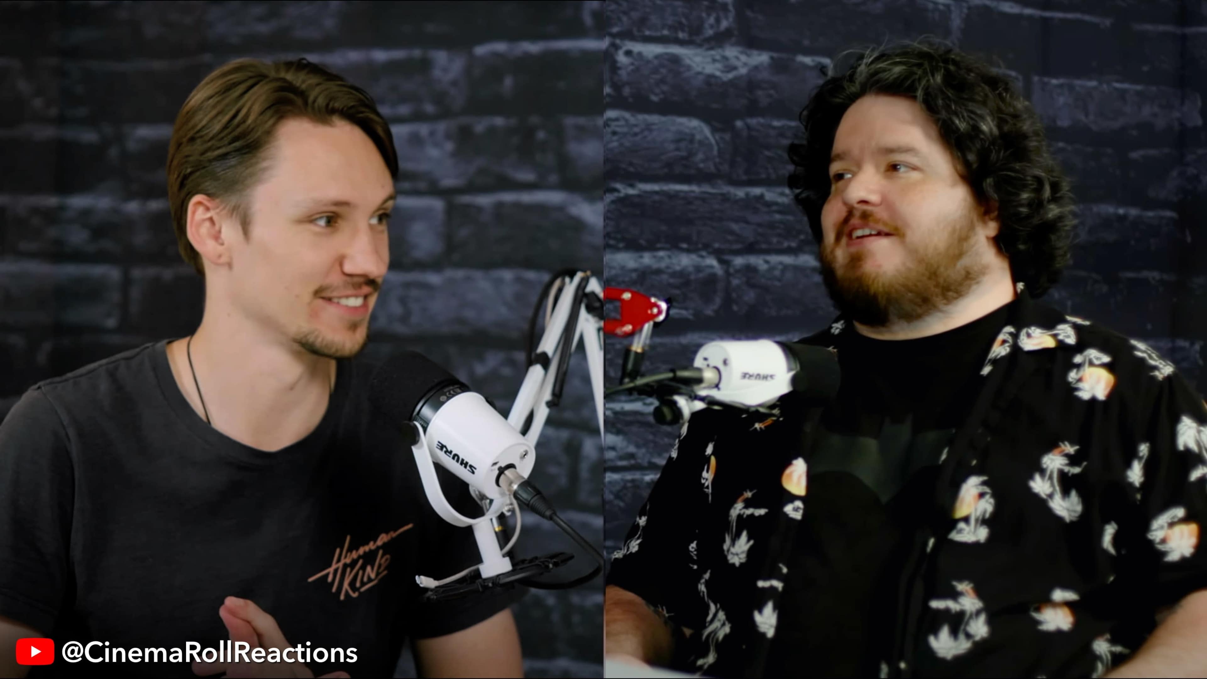 Two men sit in front of a dark brick wall, discussing while microphones are positioned in front of them.