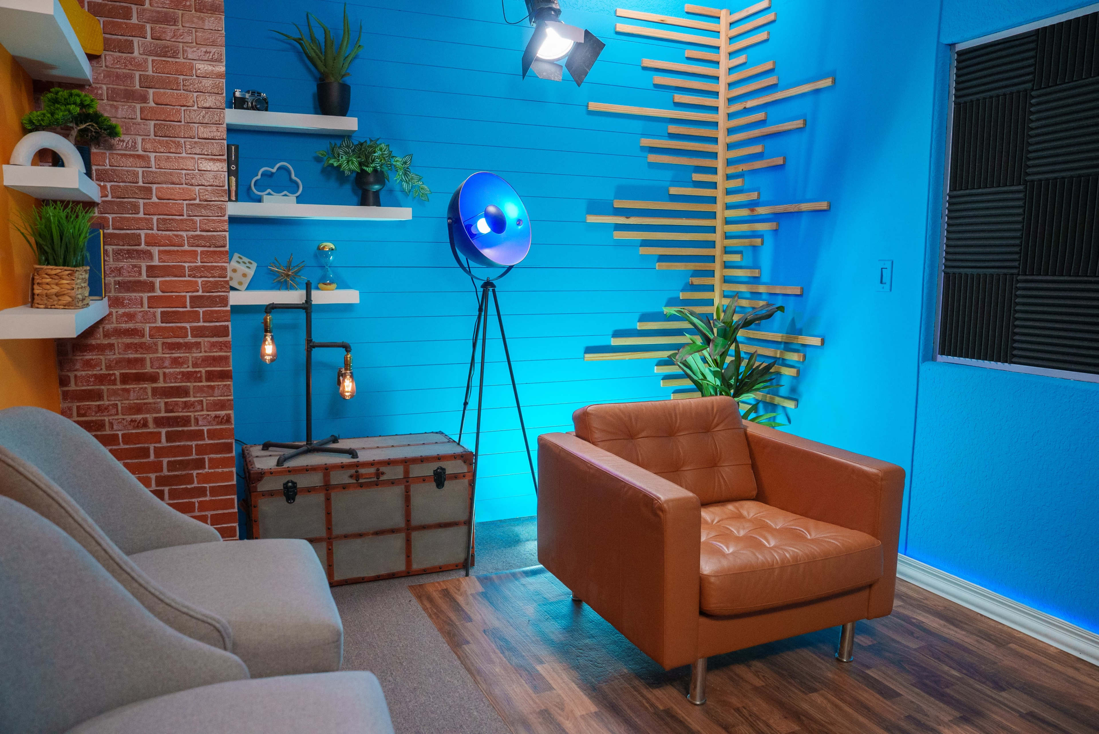 The image shows a brightly colored room featuring a blue wall with wooden accents, a brown leather armchair, gray seating, a vintage trunk, and decorative plants.