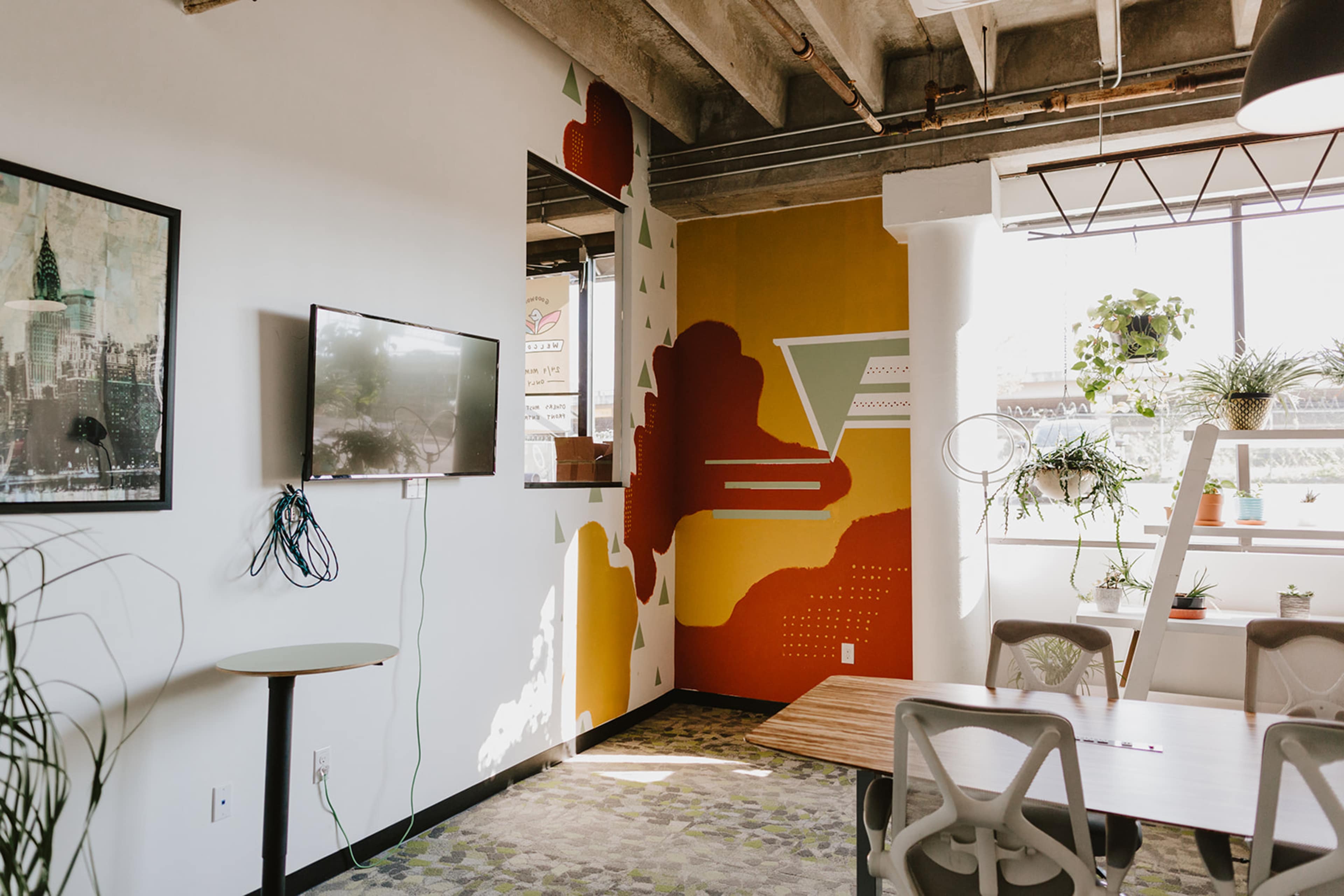 A modern office space with a TV mounted on the wall, a colorful mural, and various plants positioned around the room.