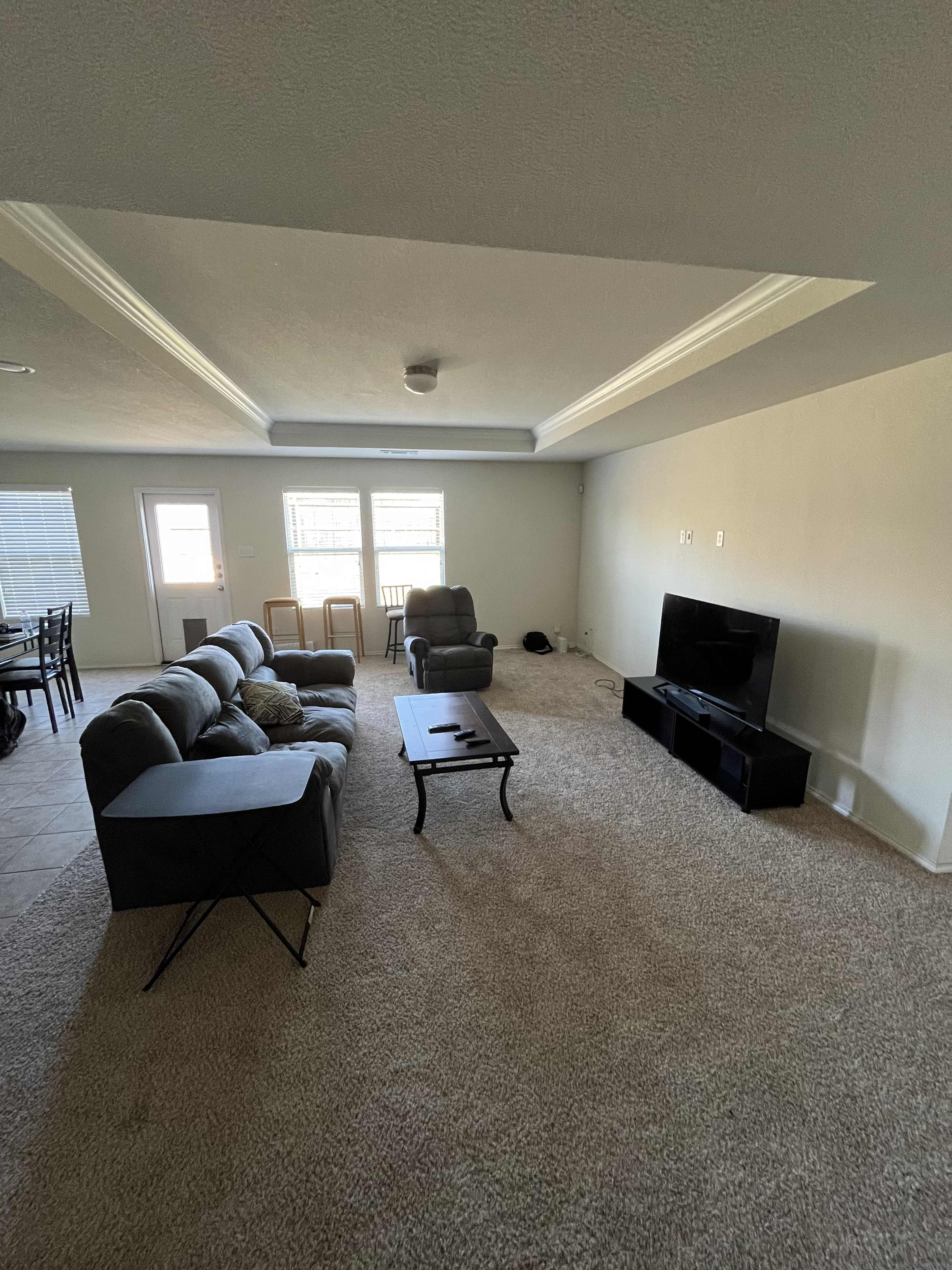The image shows a living room with a gray sectional sofa, a coffee table, a television stand, and a dining area visible in the background.