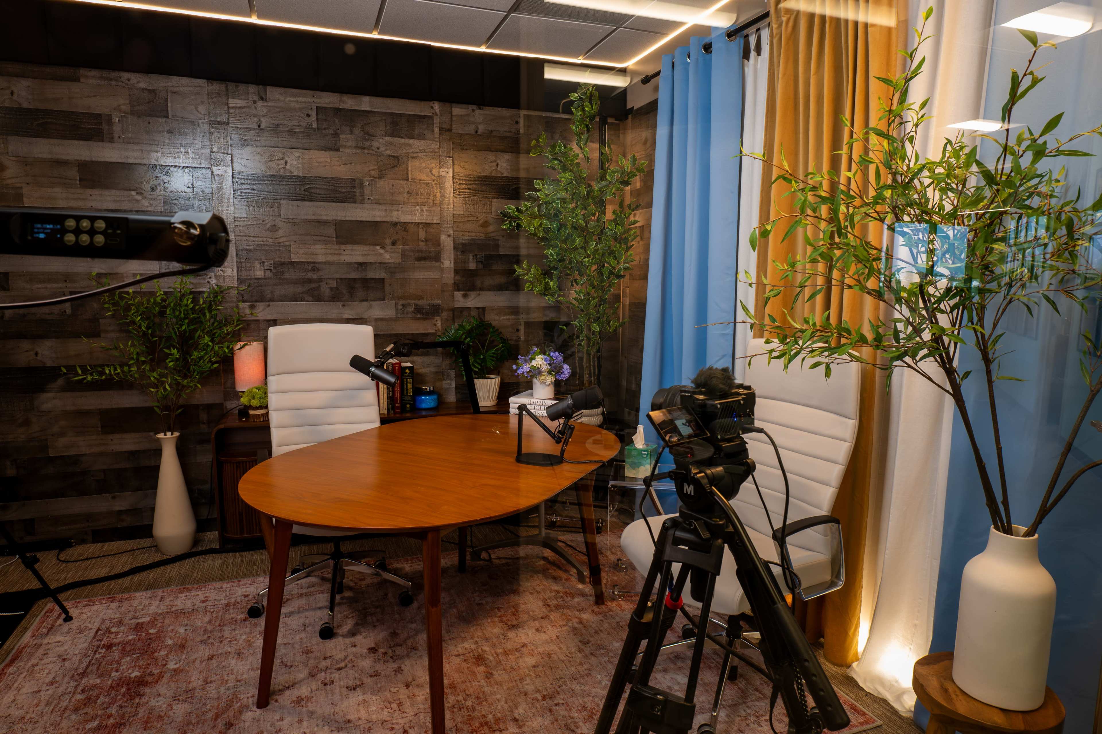 The image shows a small studio space with a wooden table surrounded by two white chairs, a camera on a tripod, and decorative plants, all under soft lighting.