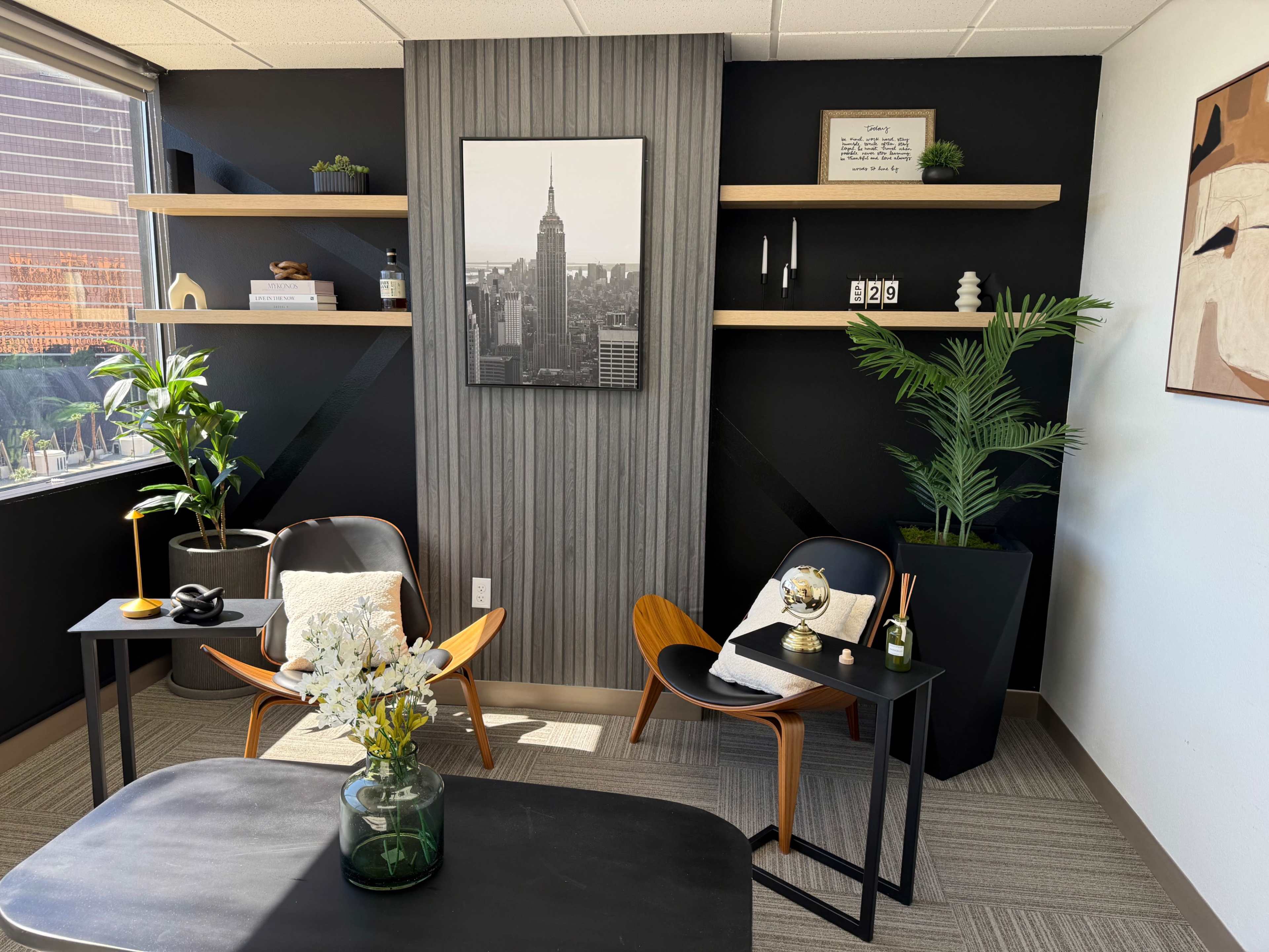 The image shows a modern office space featuring two chairs, a small table, decorative shelves on the wall, and plants.