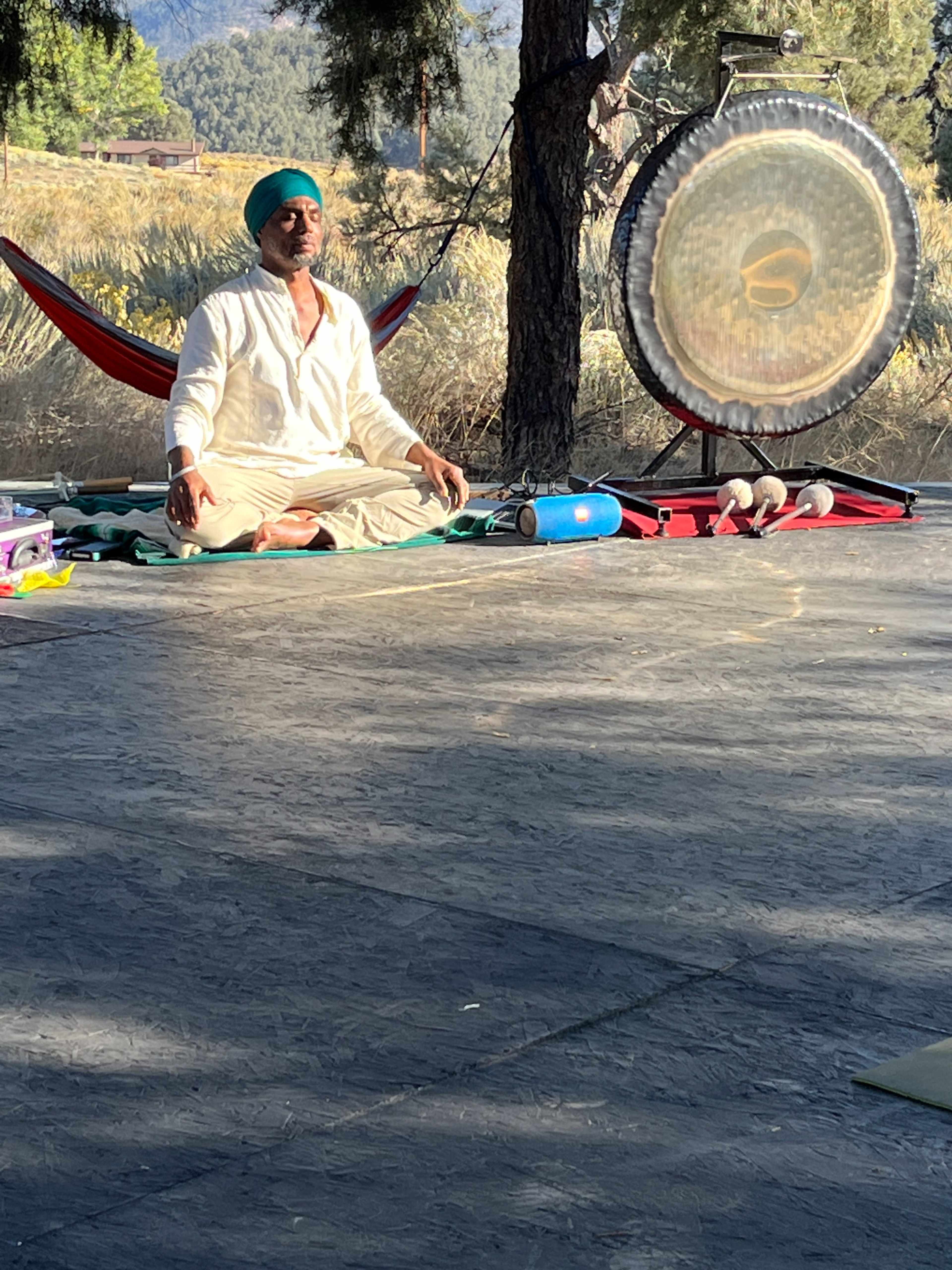 A person sits cross-legged on a wooden platform near a large gong, surrounded by trees and a hammock in the background.