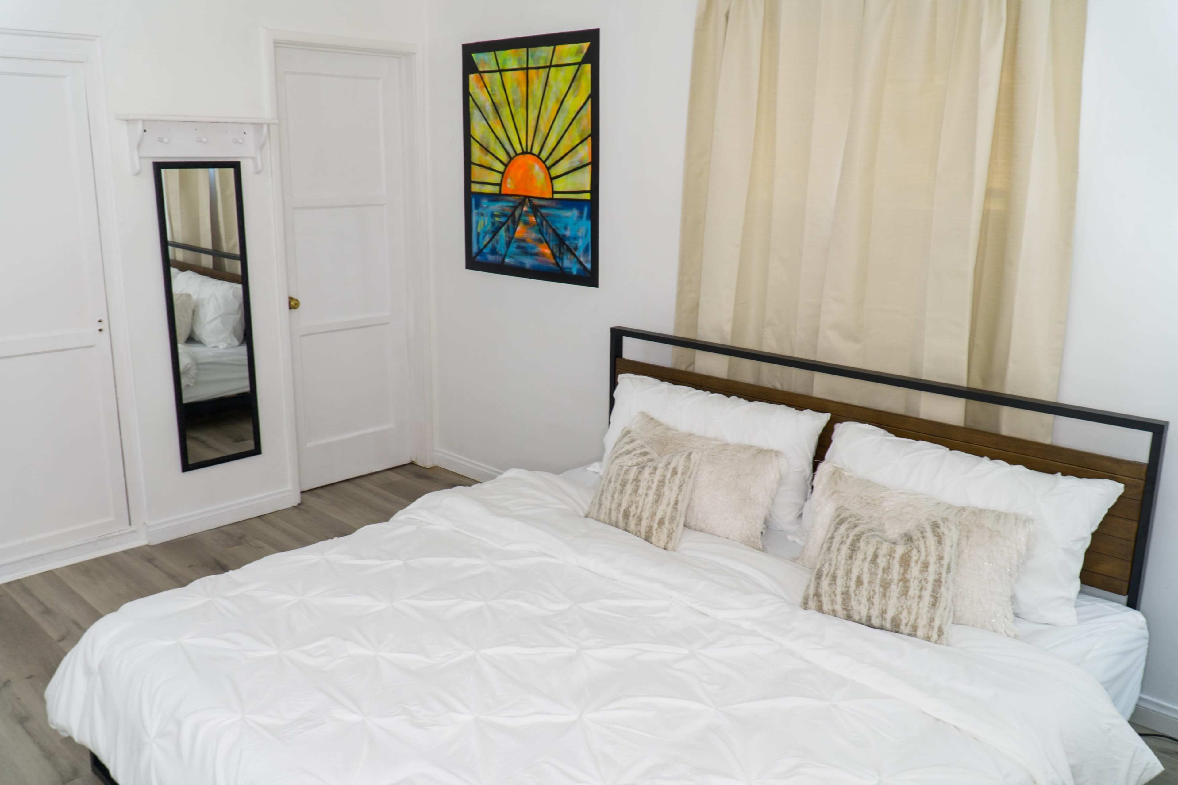 A neatly arranged bedroom features a bed with white bedding, a large window with cream curtains, a colorful sun painting on the wall, and a mirrored closet door.