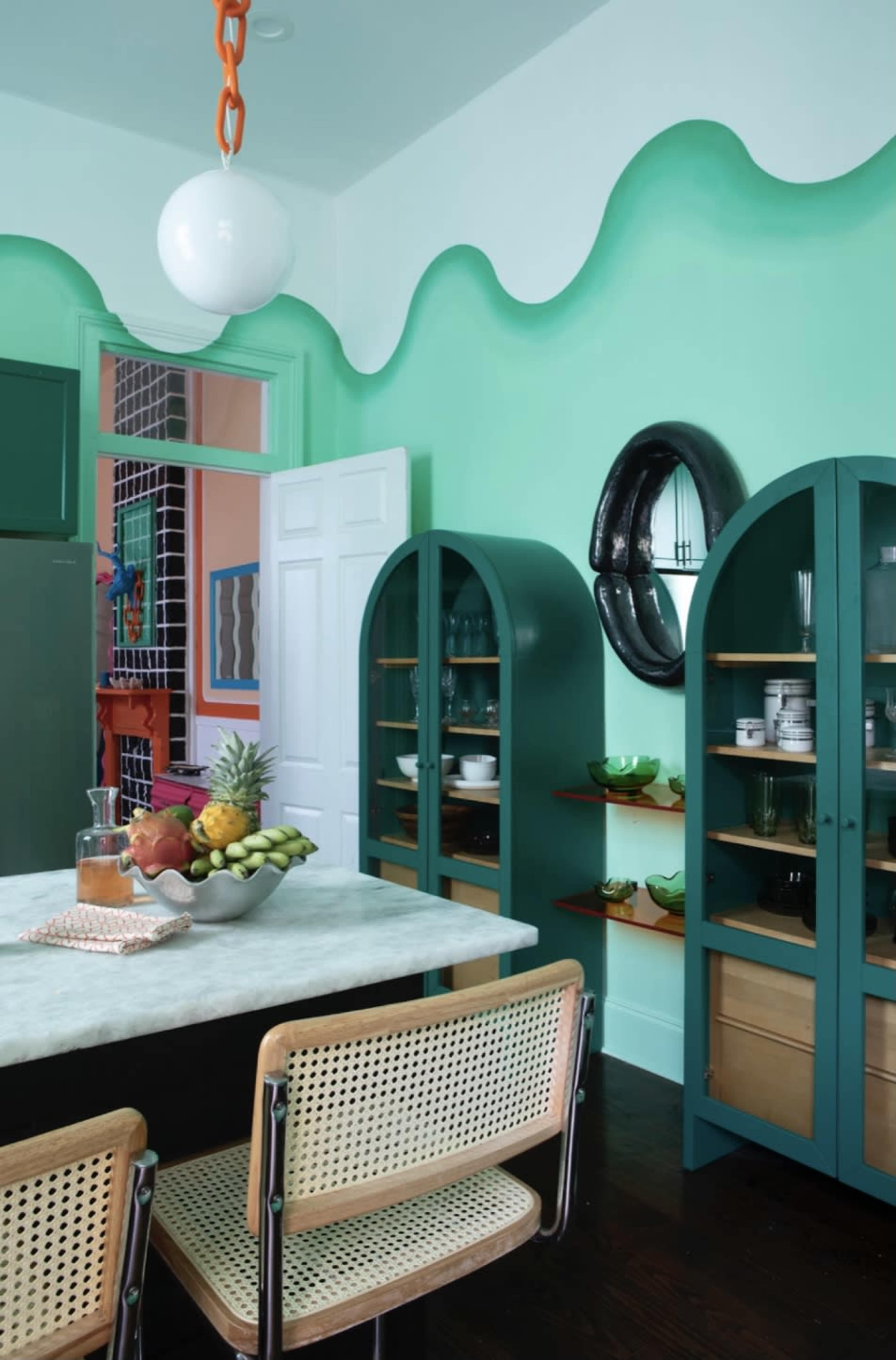 The kitchen features green wave-patterned walls, a marble table, and wooden cabinets displaying various dishware and fresh fruits.