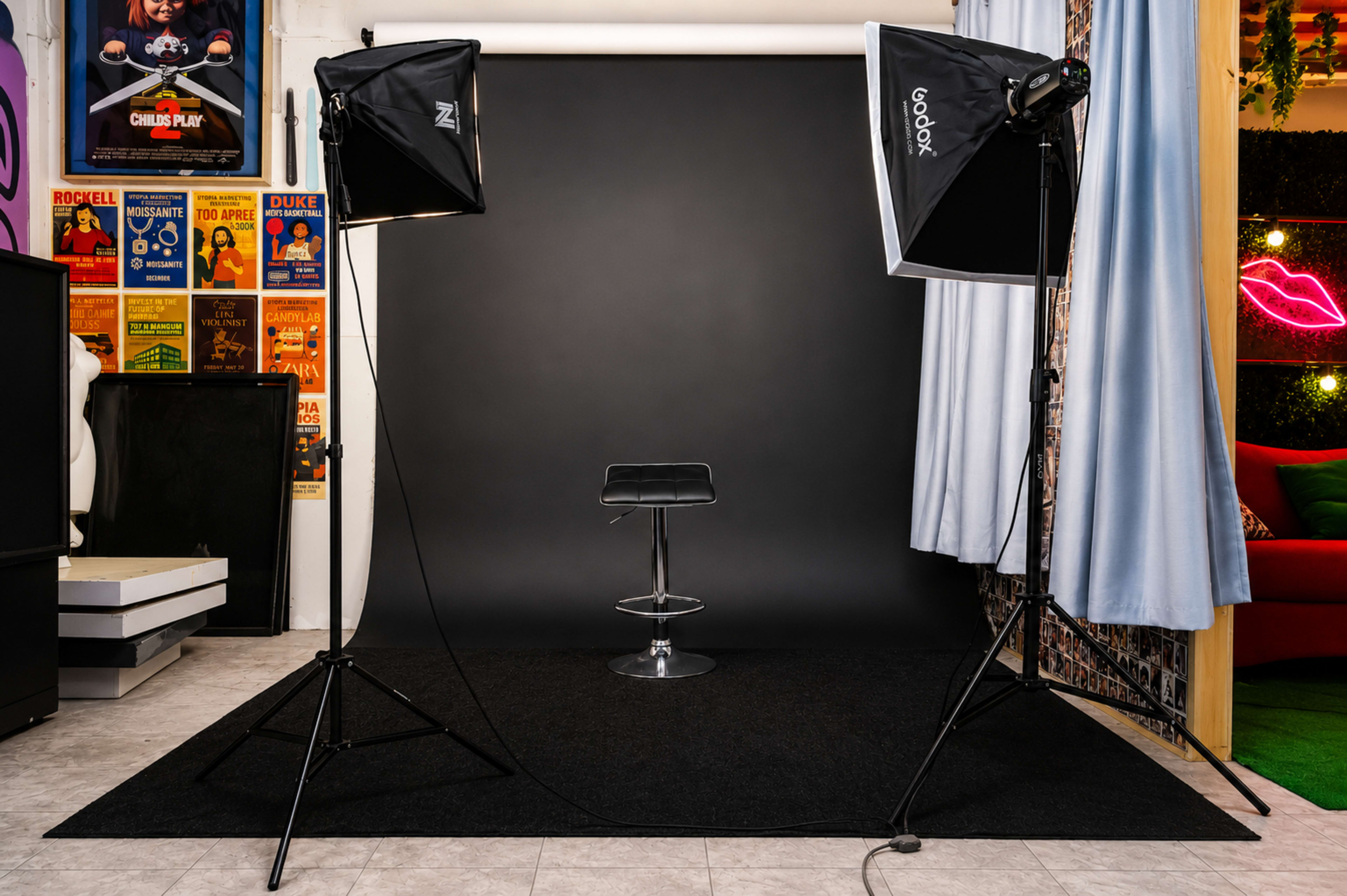 The image shows a photography studio setup with a black backdrop, a stool in the center, and two softbox lights positioned on either side.