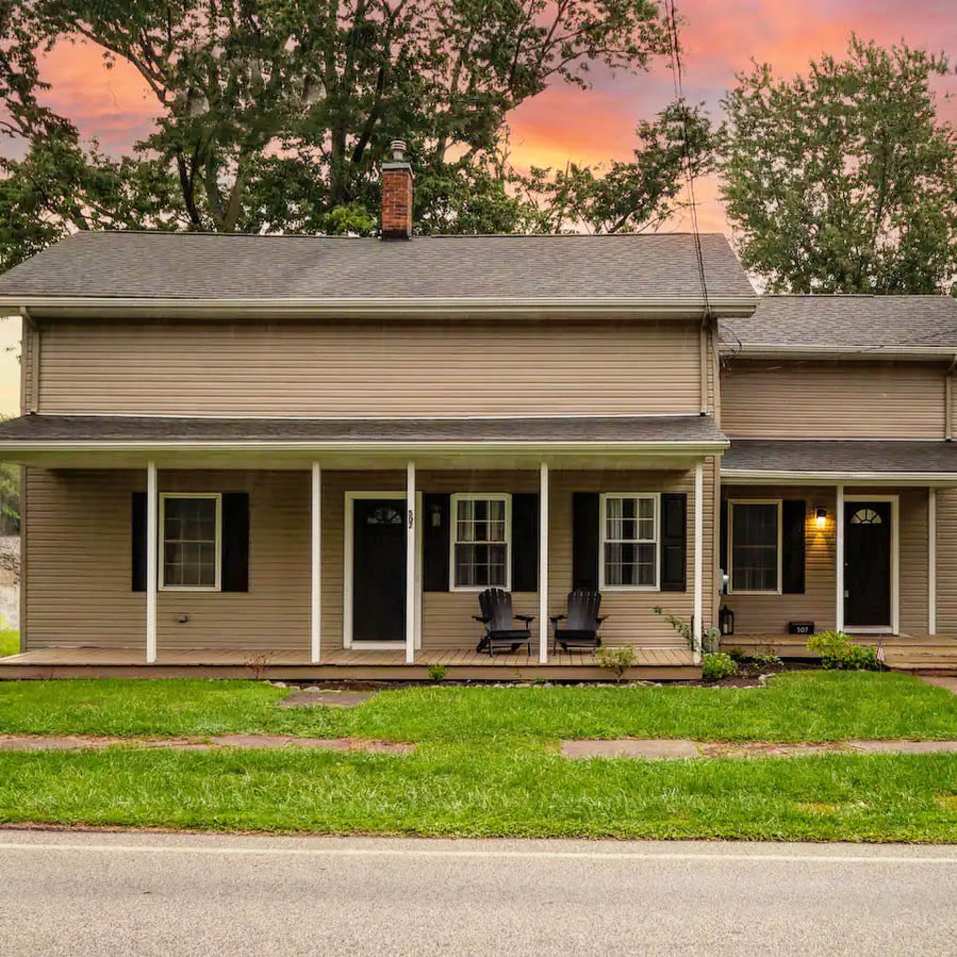 A two-story house with a front porch, two black chairs, and a well-maintained lawn, set against a colorful sunset sky.