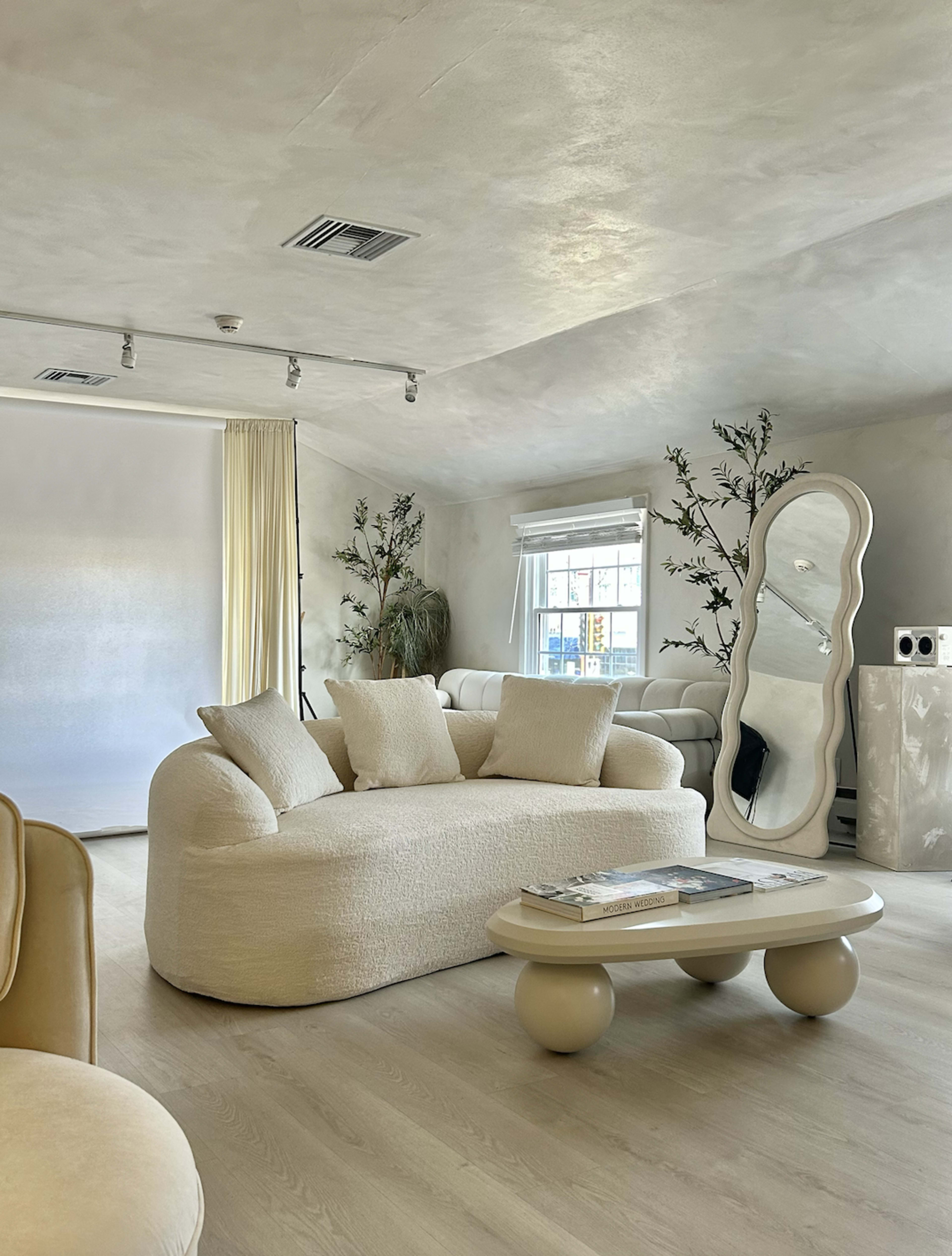 The image shows a modern living room with a curved white sofa, a round coffee table, and large mirrors, all set against light-colored walls.