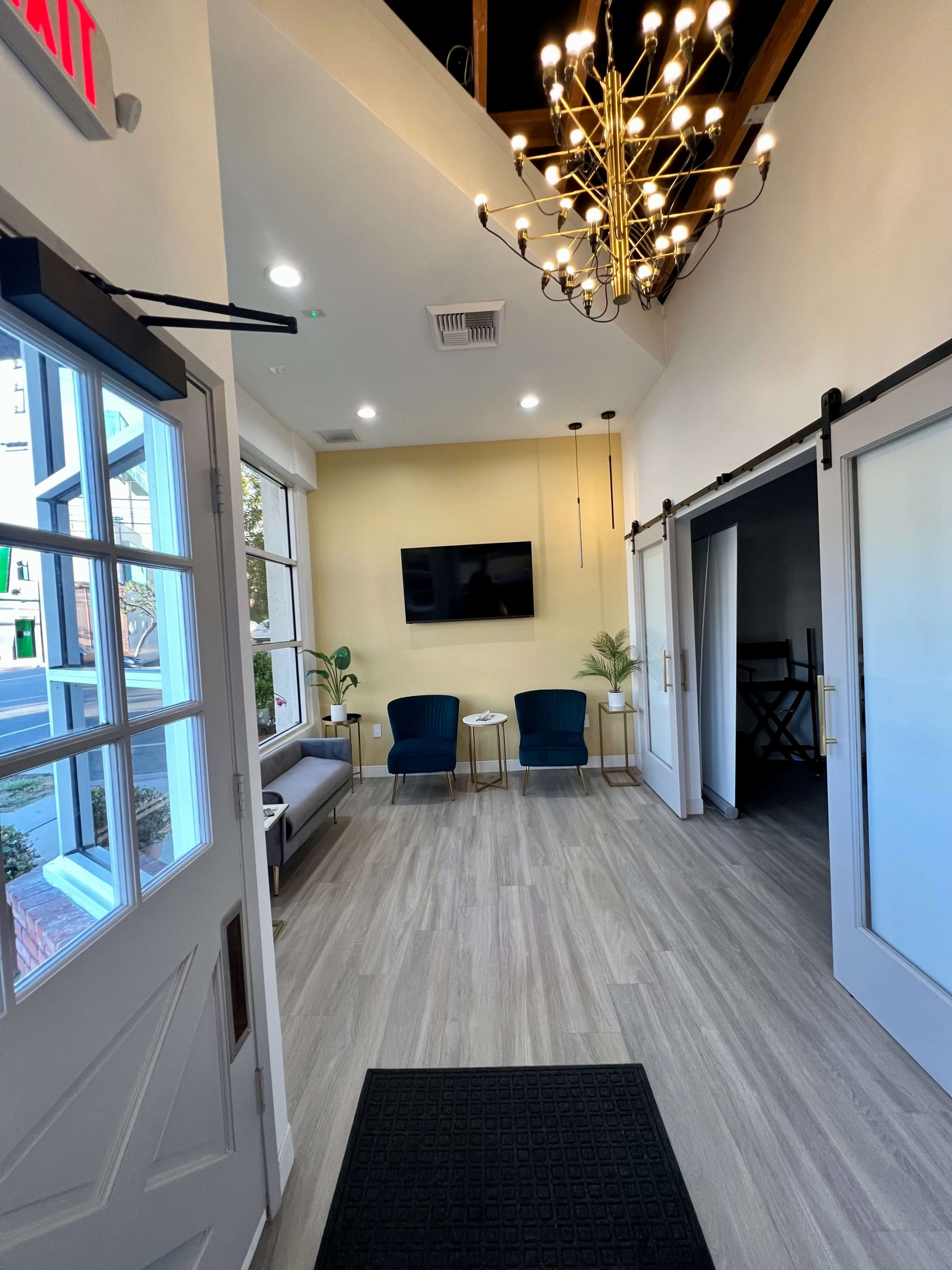 A well-lit entryway with a modern chandelier, two chairs, a television on the wall, and sliding doors leading to another room.