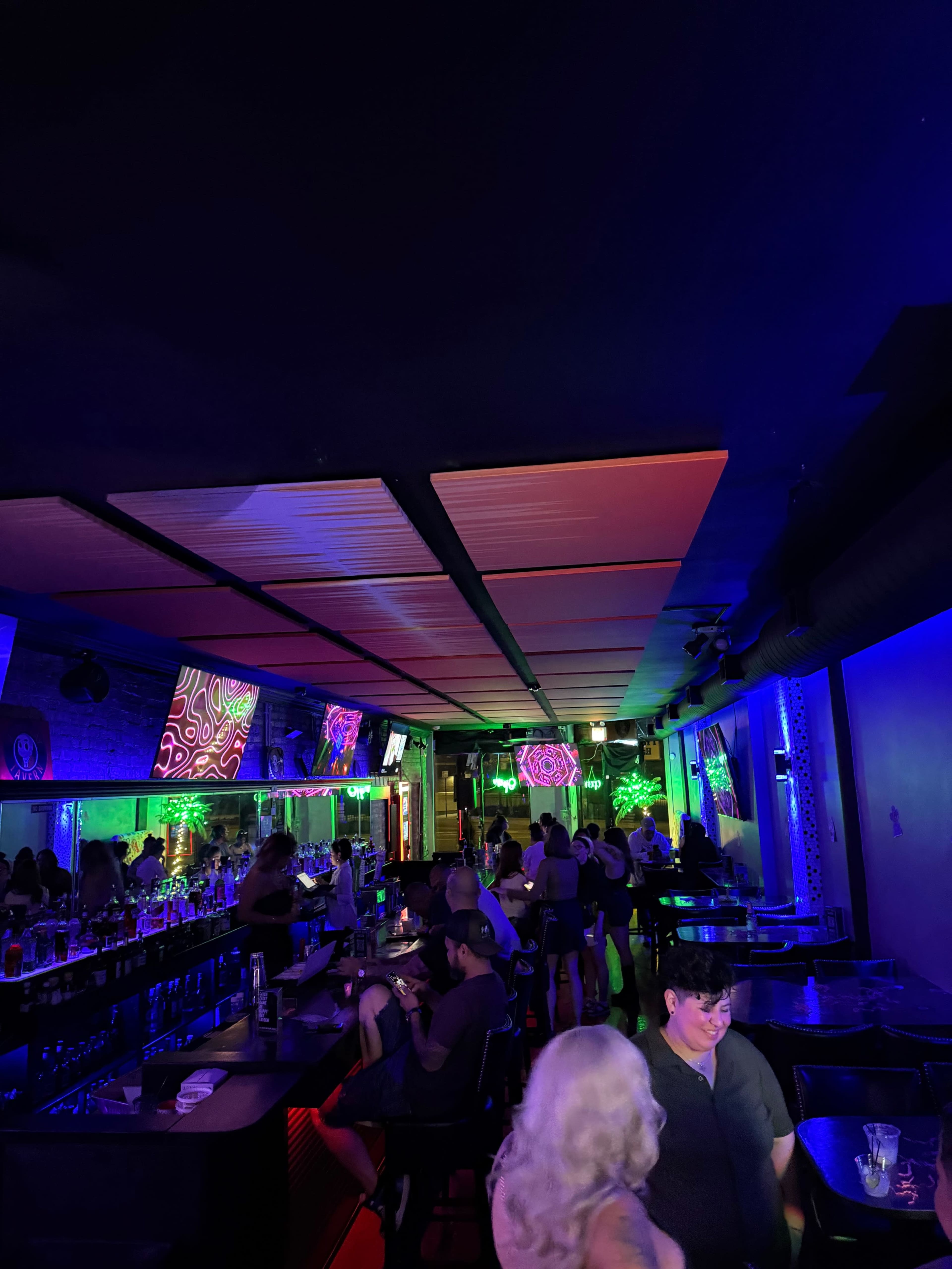 The image shows a vibrant bar scene with colorful lighting, featuring several patrons seated and standing in a lively atmosphere.