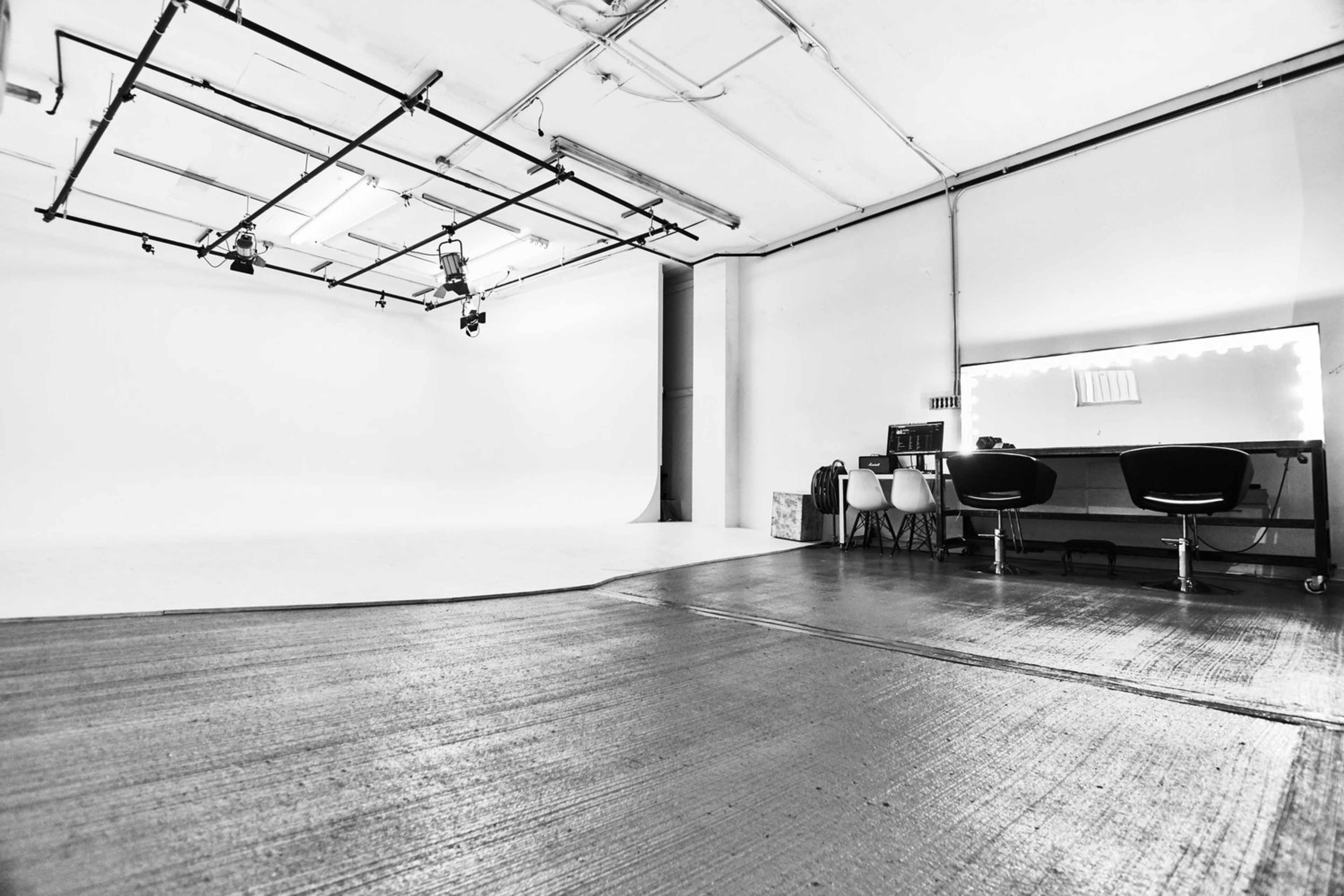 A minimalist photography studio with a white backdrop, overhead lighting, and a row of chairs beside a vanity mirror.