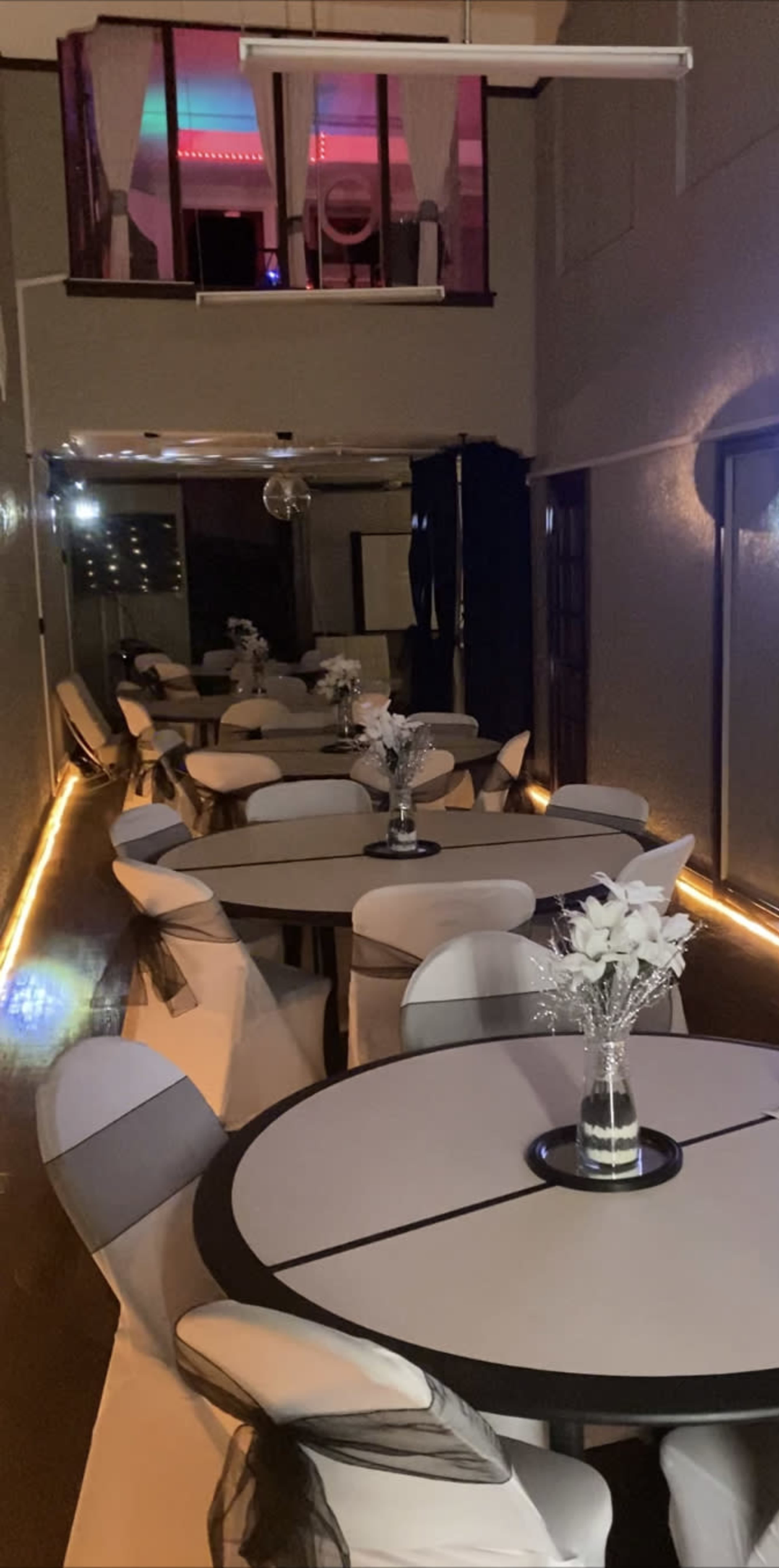 A narrow dining space features several round tables adorned with floral centerpieces and low lighting along the floor.