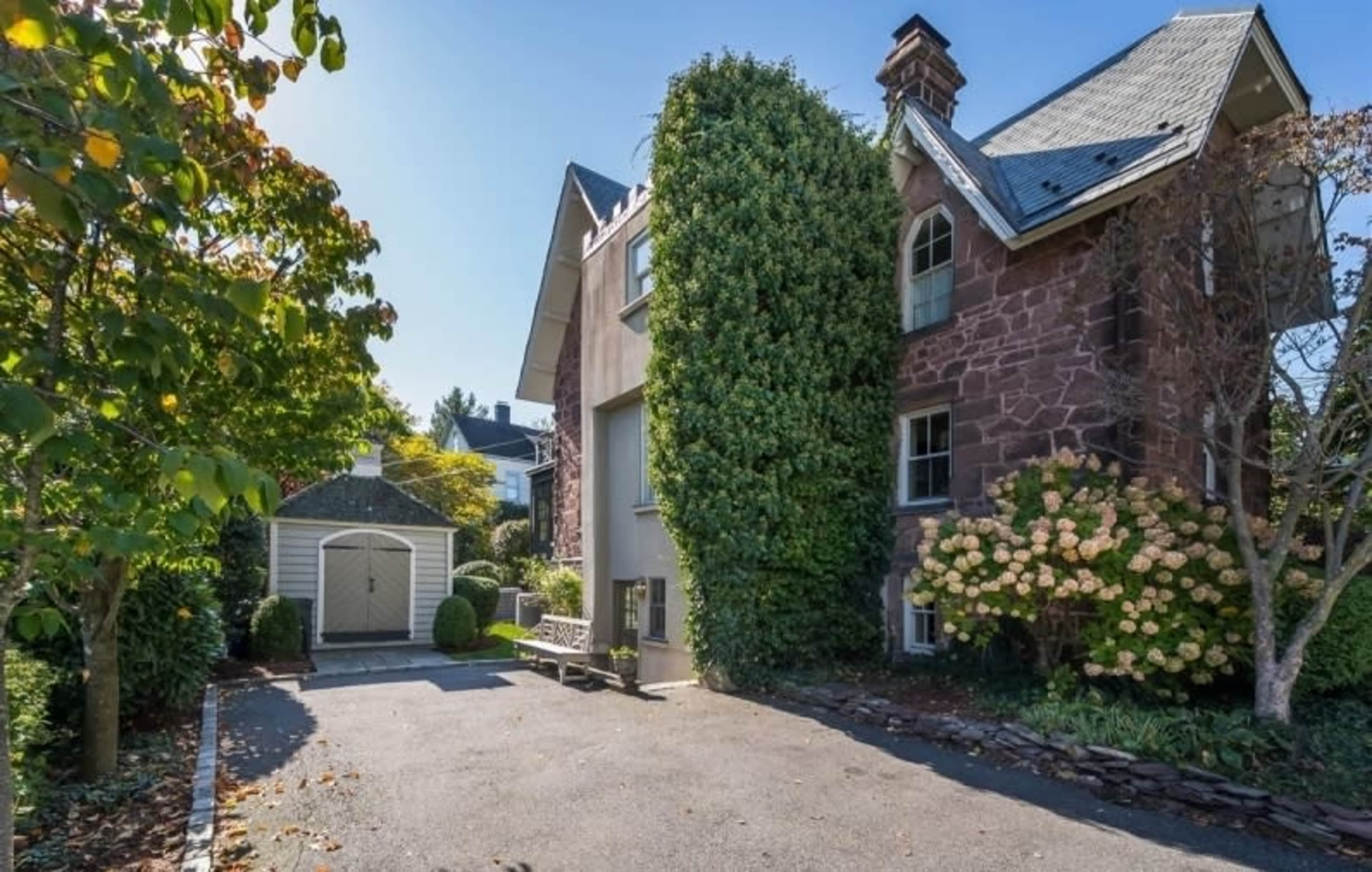 The image shows a stone house with a tall green vine covering part of its facade, a gravel driveway, and a small garage on the side.