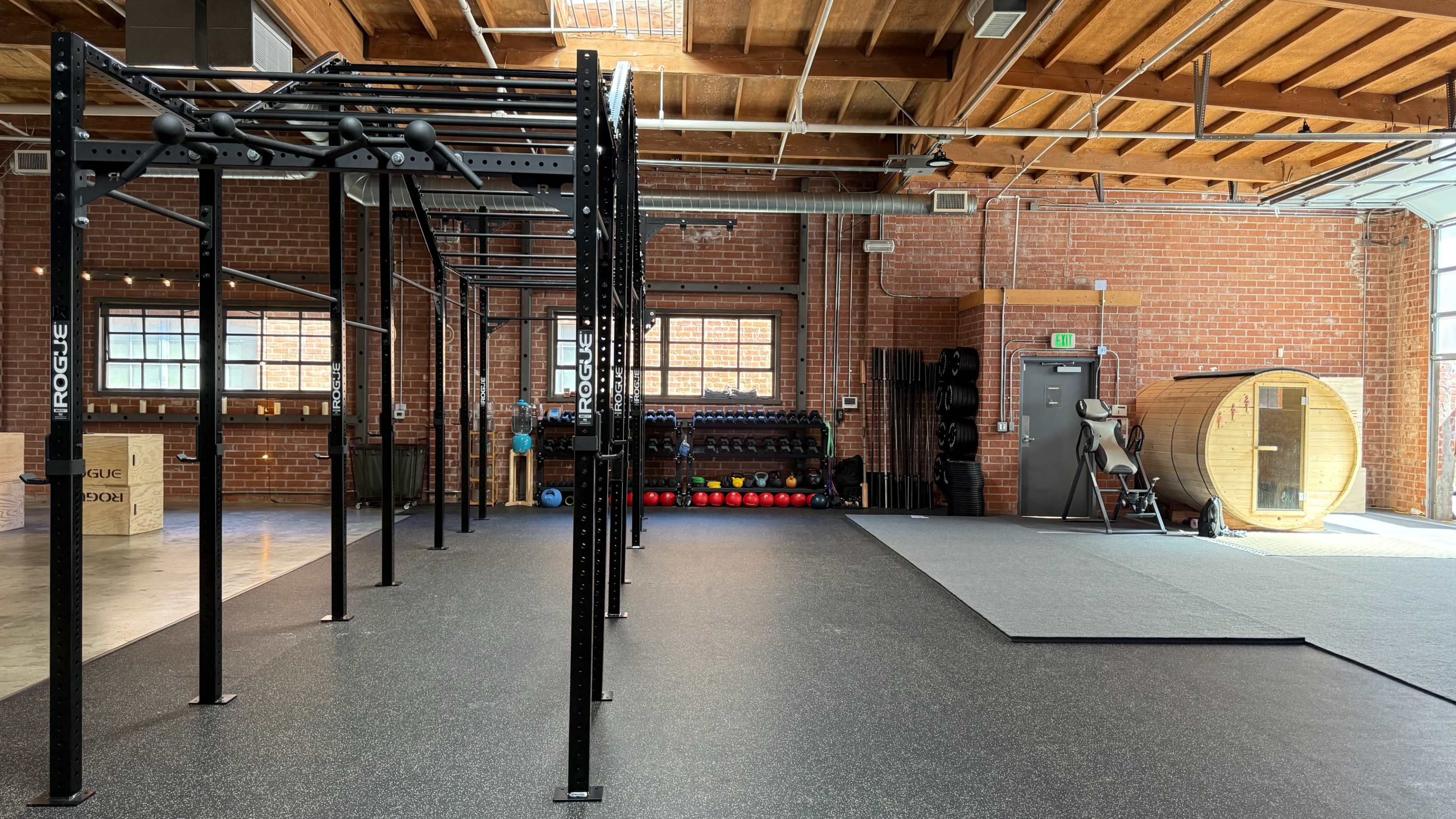 The image shows a spacious gym interior with black exercise equipment, including a pull-up structure, free weights, and a circular wooden sauna, under wooden beams and brick walls.