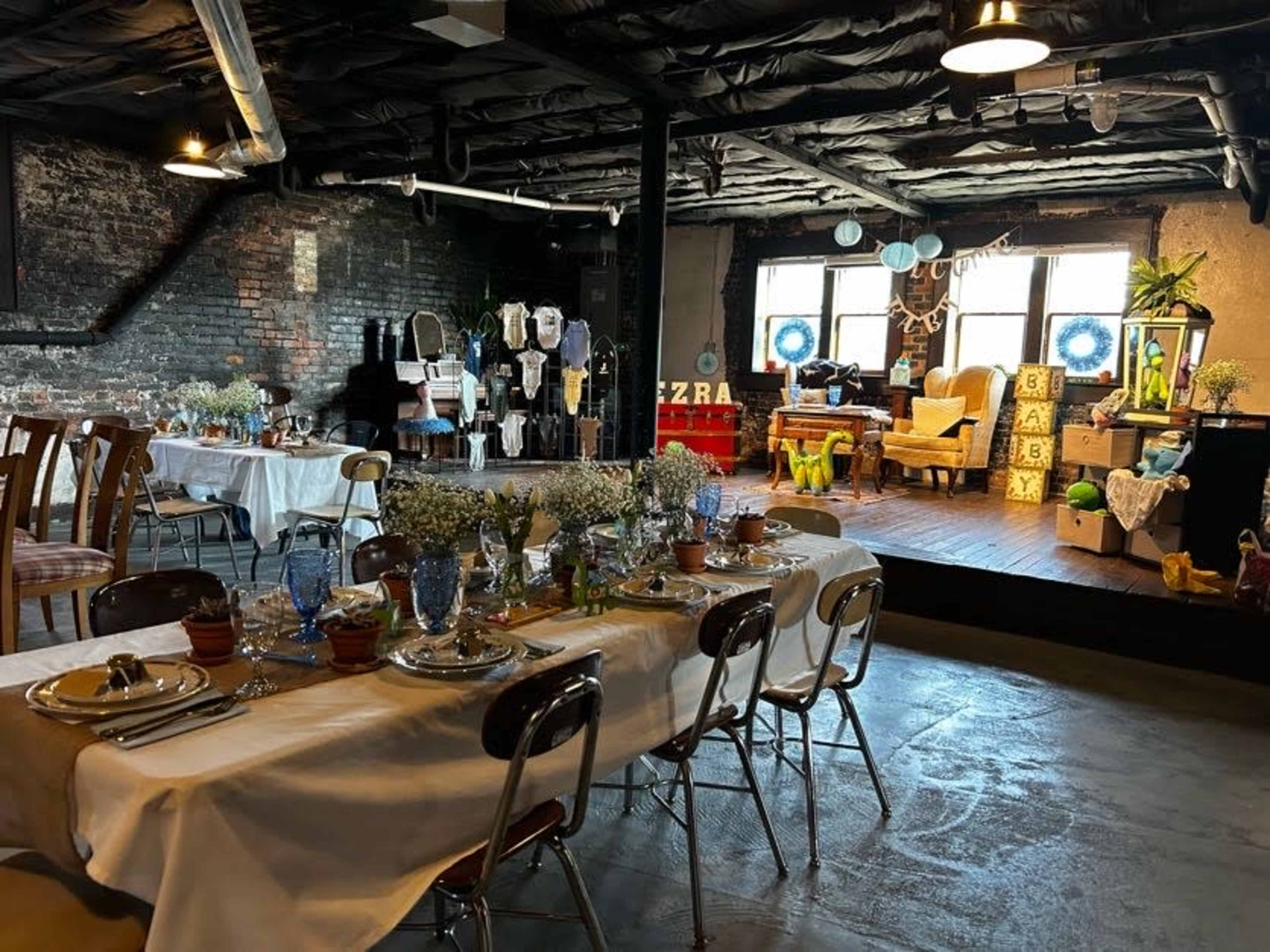 The image shows a spacious industrial-style room set up for an event, featuring a long dining table with place settings and a decorative area with chairs and various display items in the background.