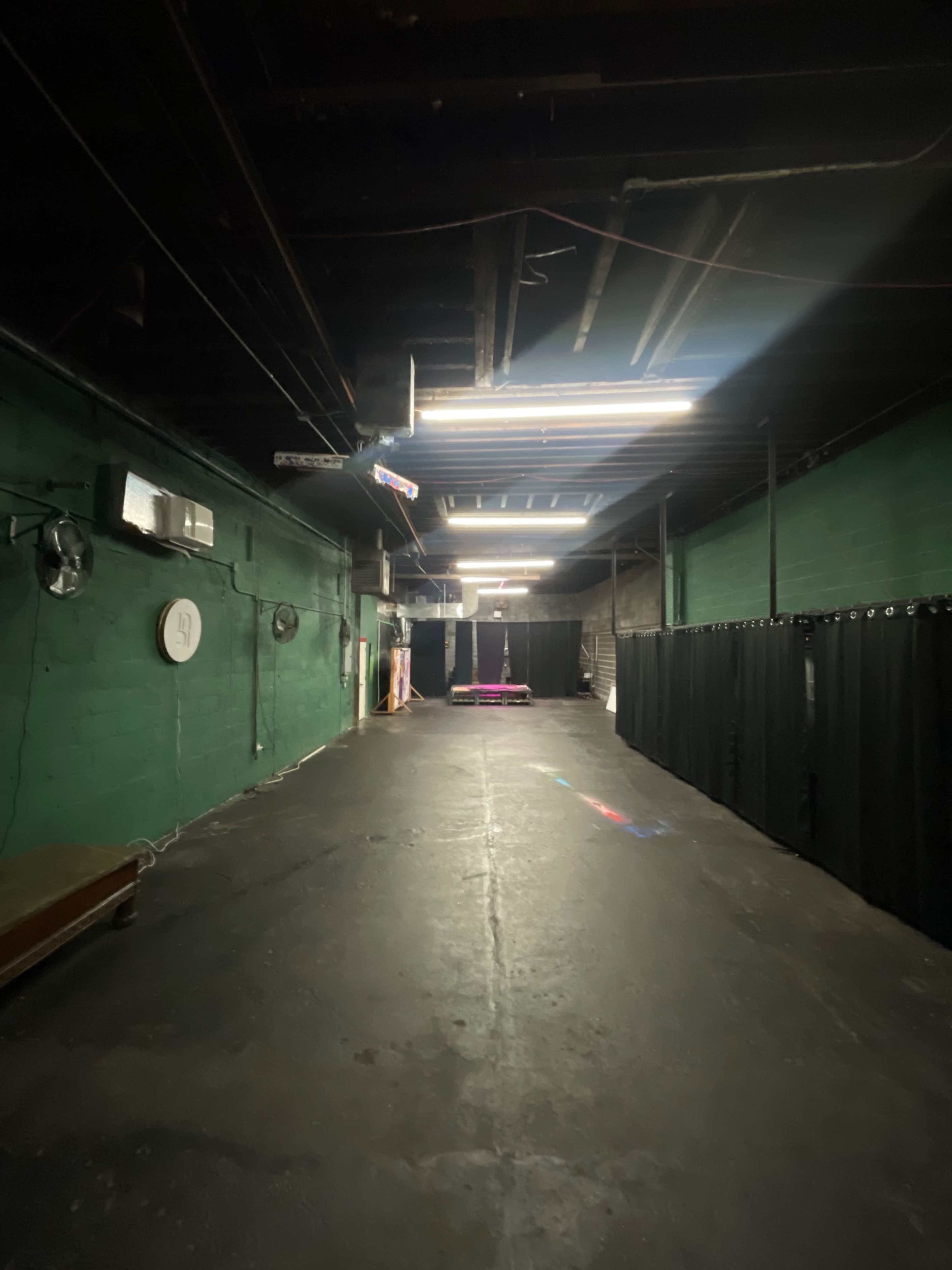 The image shows a dimly lit, empty corridor in a green-painted building with black curtains on one side and a stage area illuminated at the far end.