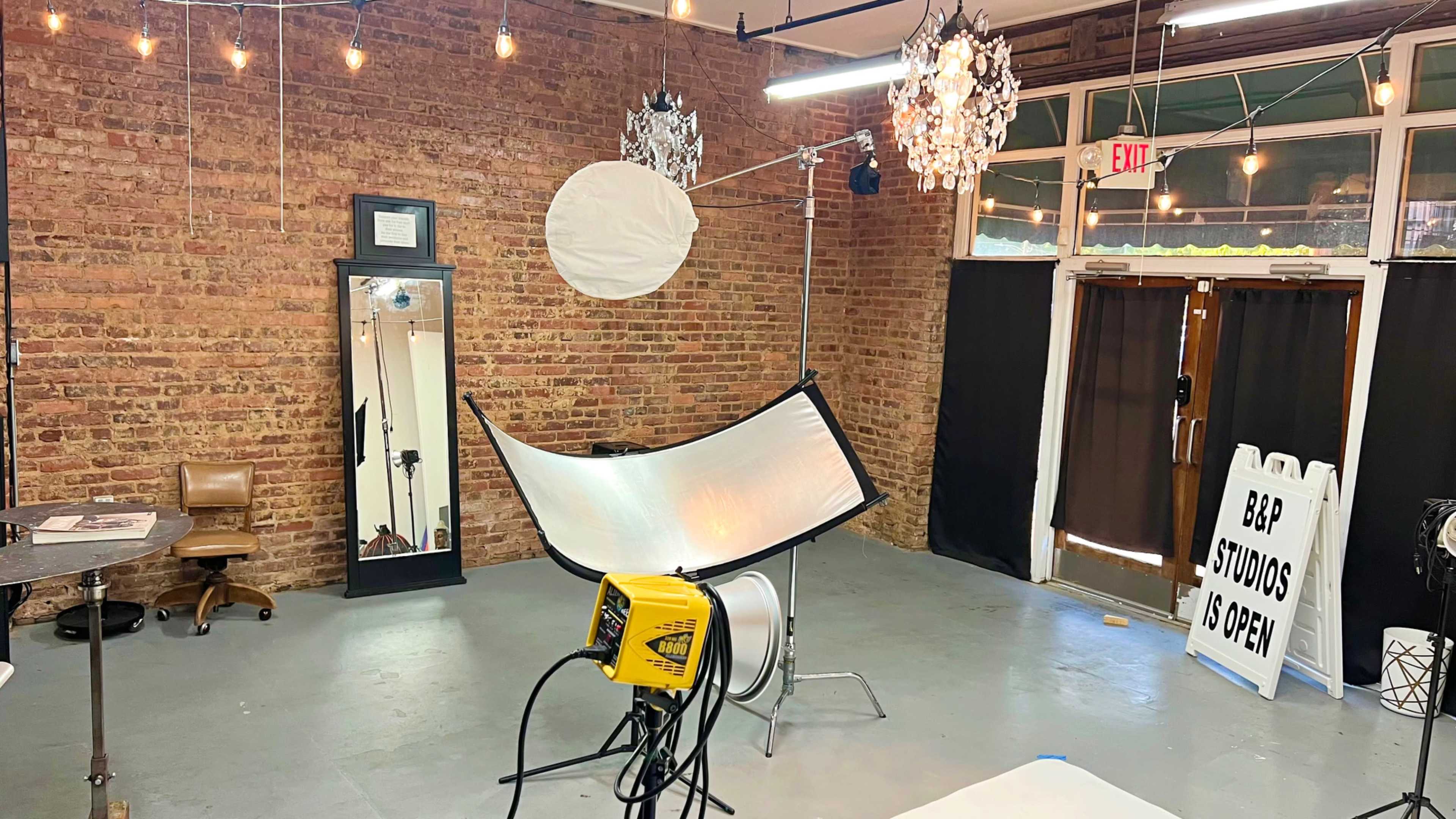 The image shows a photography studio with brick walls, lighting equipment, a large reflector, and a mirror, featuring a sign indicating that the studio is open.