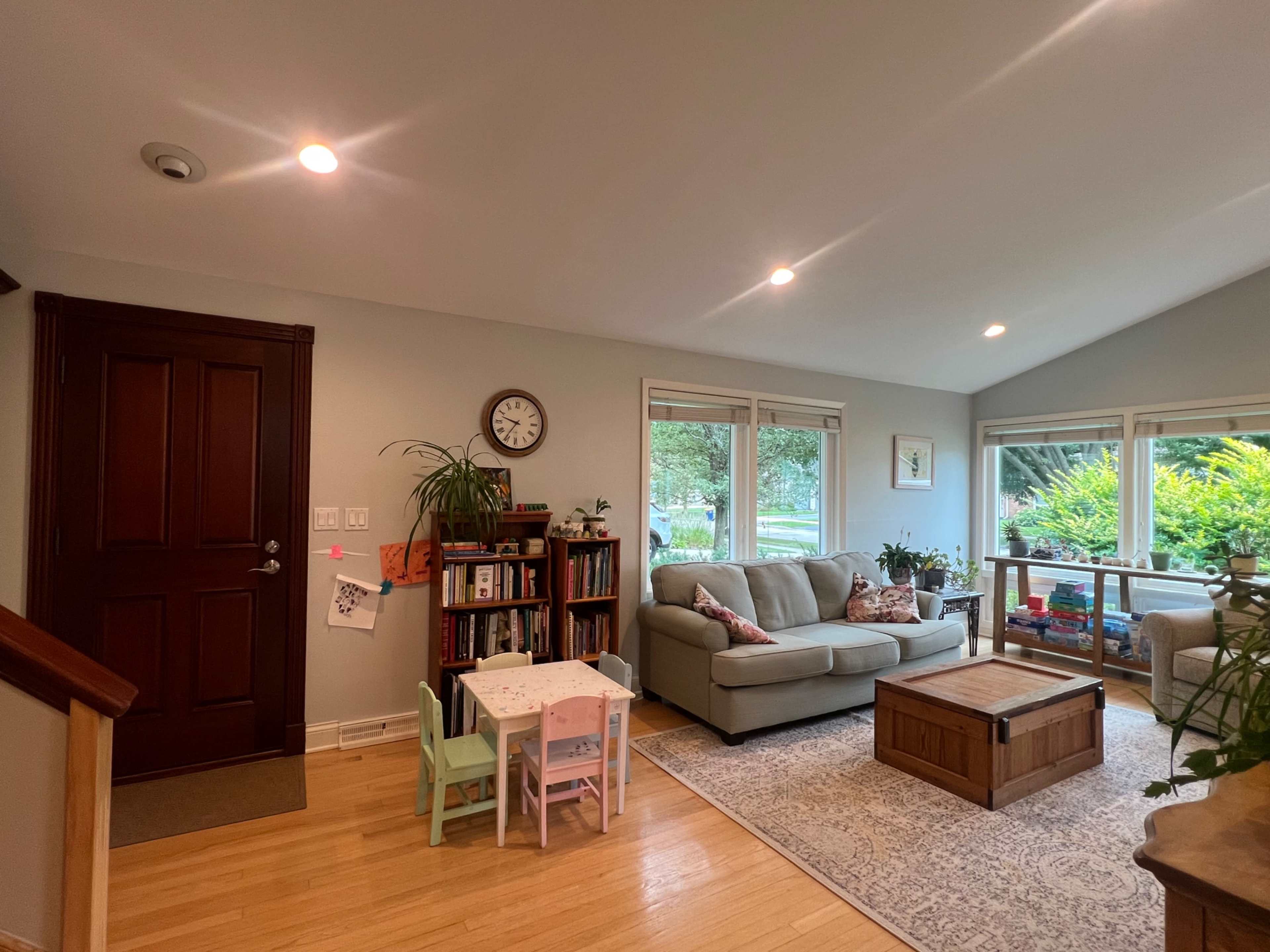 A cozy living room features a light-colored sofa, a wooden coffee table, a small children's table with chairs, and bookshelves against the wall, with large windows showing a green outdoor view.