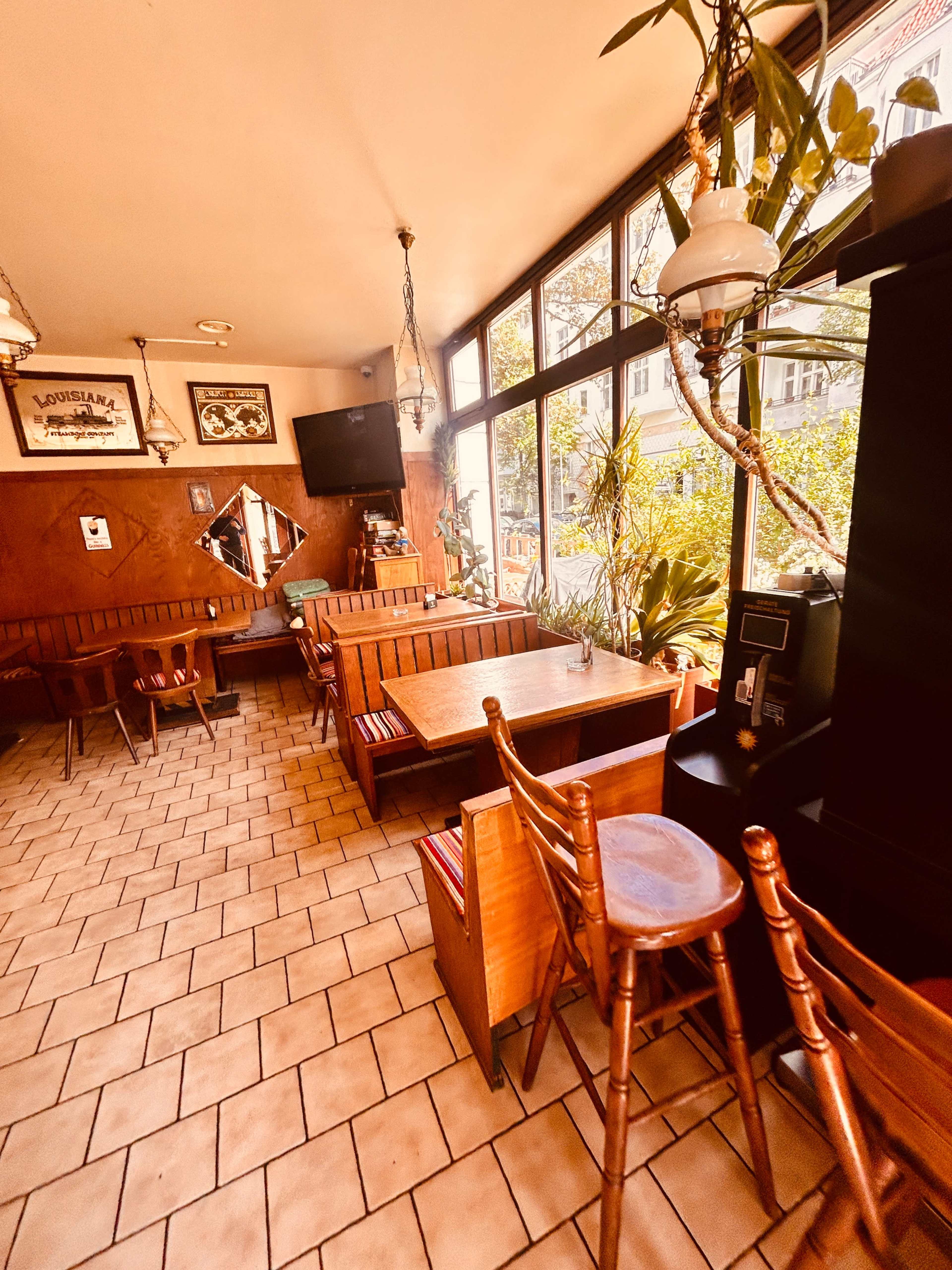 The image shows a cozy restaurant interior with wooden tables and chairs, large windows, and plant decorations.