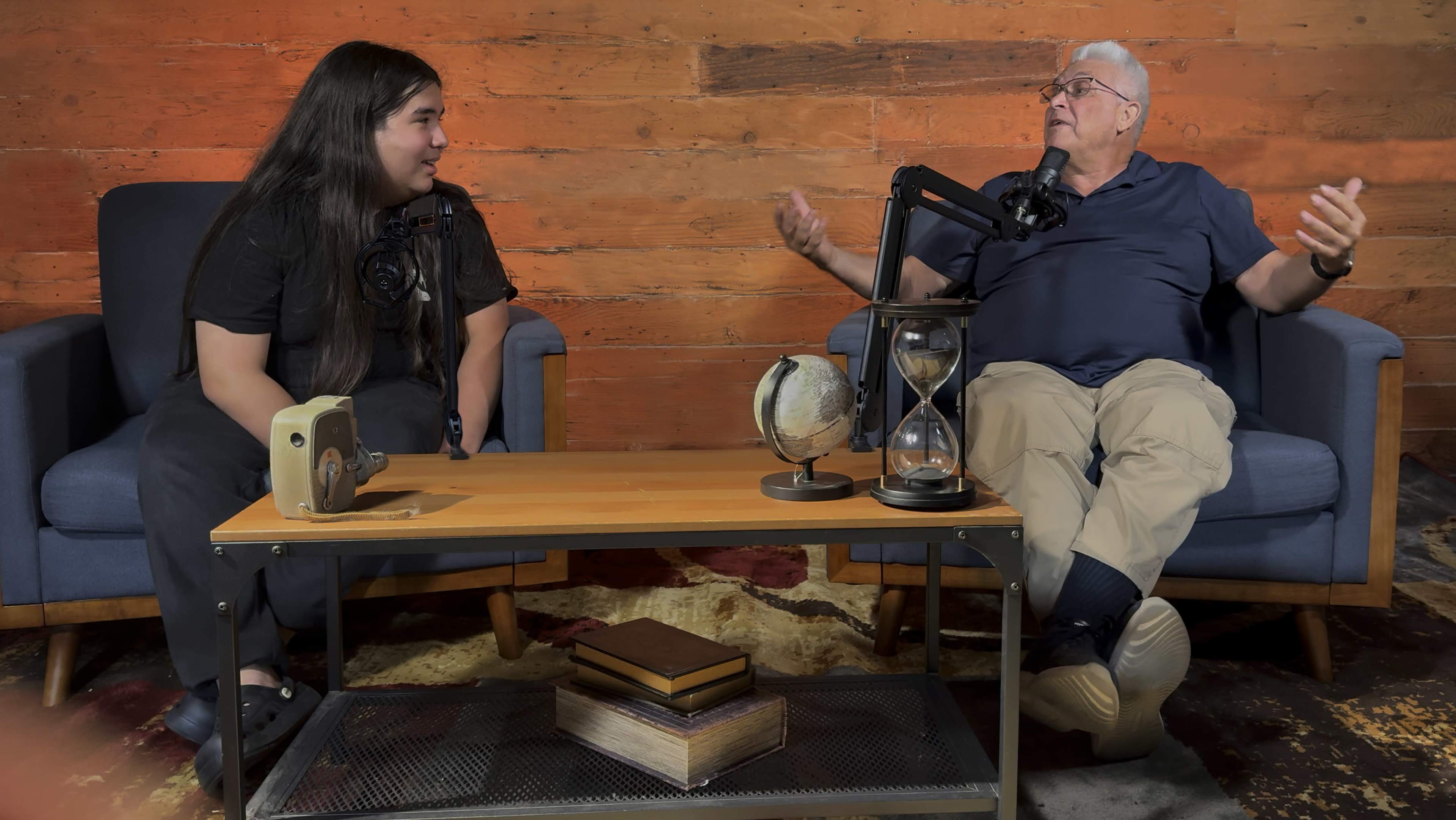 A young person and an older man are engaged in conversation while seated on blue armchairs, with microphones positioned in front of them on a wooden table.