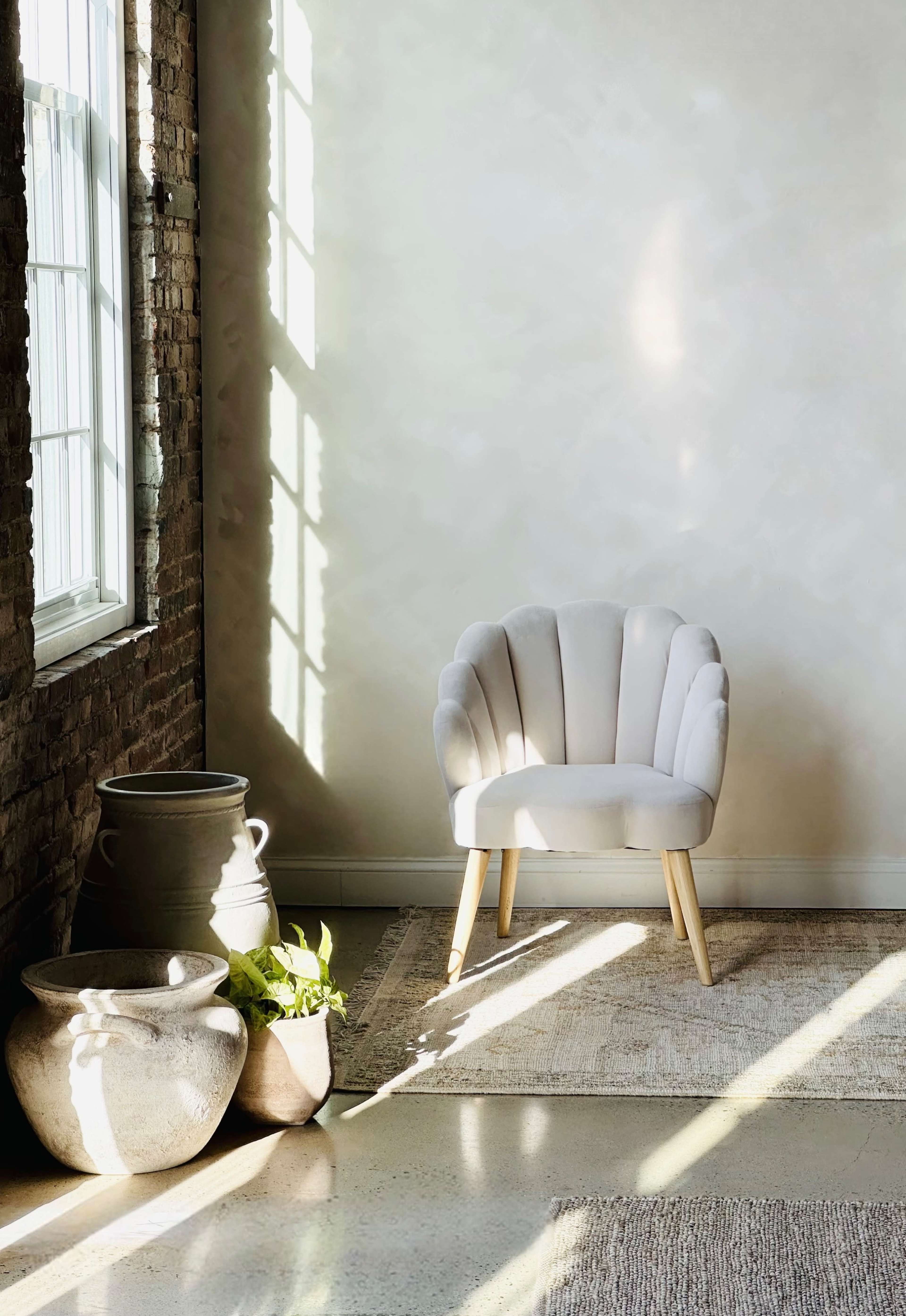A light-colored chair with a scalloped design is positioned next to potted plants and pottery in a sunlit corner near a large window.