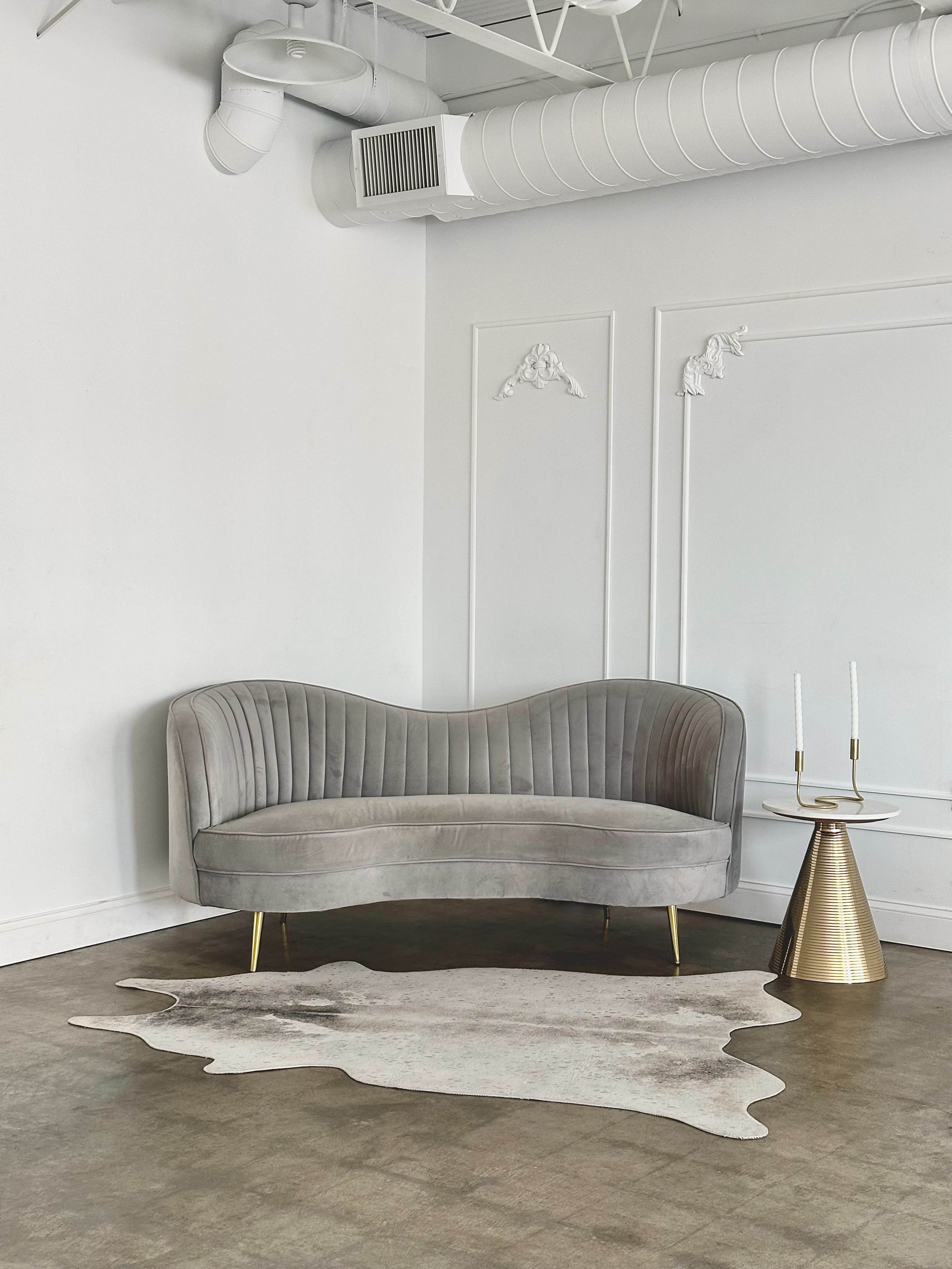 A modern gray curved sofa is positioned next to a metallic side table with two candles on a light-colored cowhide rug.