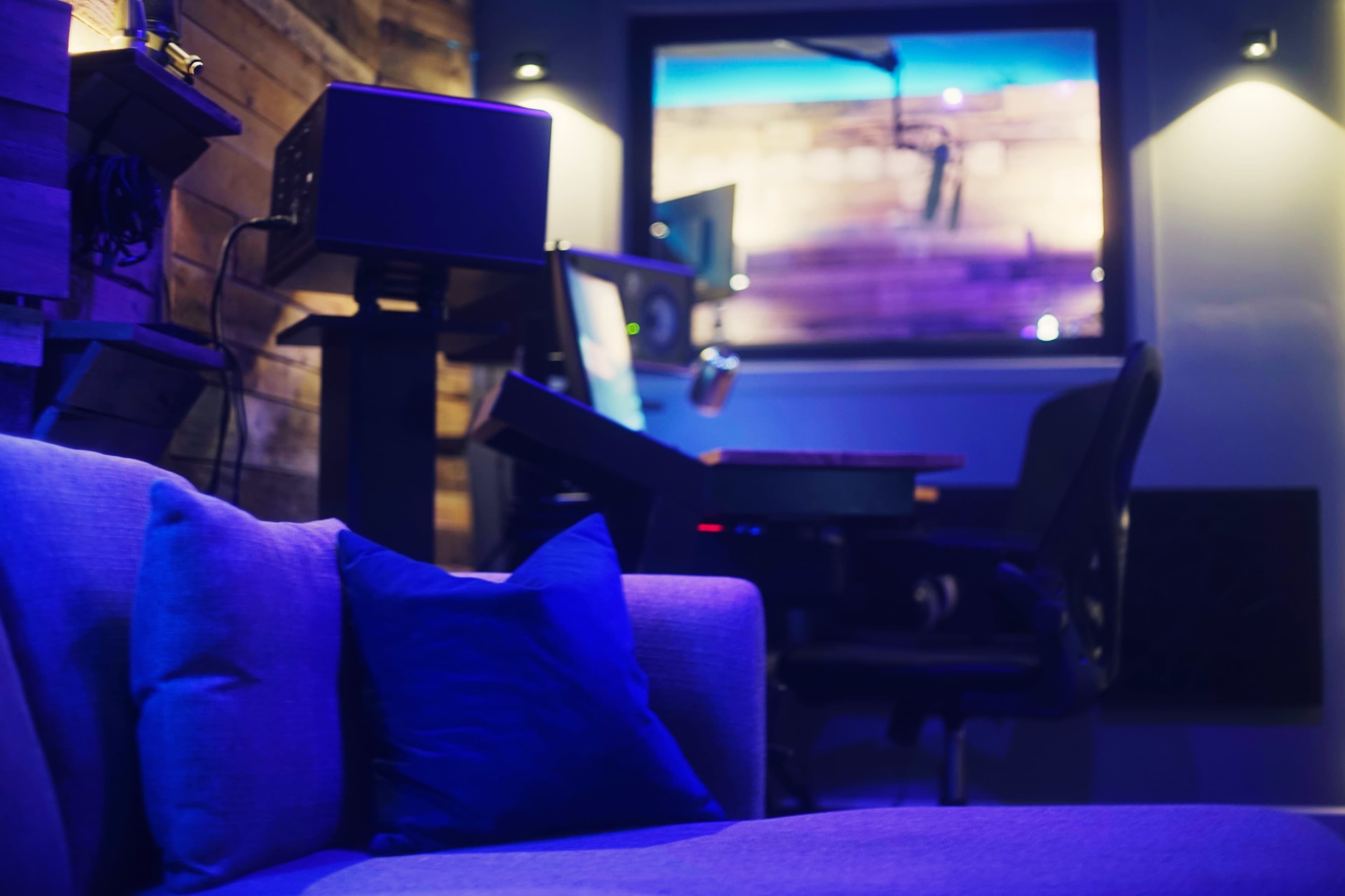 A modern workspace is visible in the background, featuring a sleek desk with equipment and a large window illuminated by purple lighting, while a blue sofa with a decorative pillow is in the foreground.