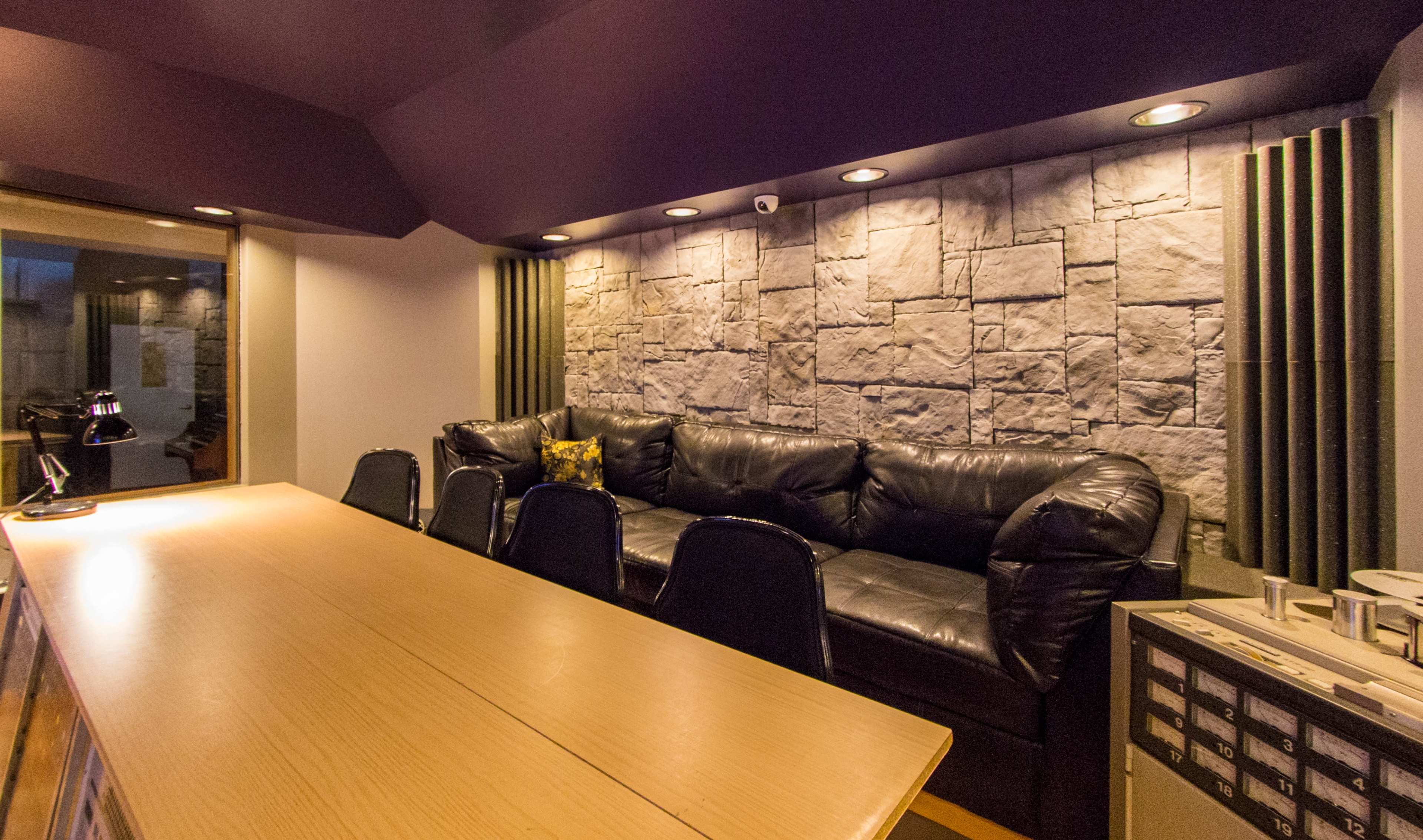 The image shows a dimly lit lounge area featuring a black leather sofa, a wooden bar counter, and a stone wall with recessed lighting.