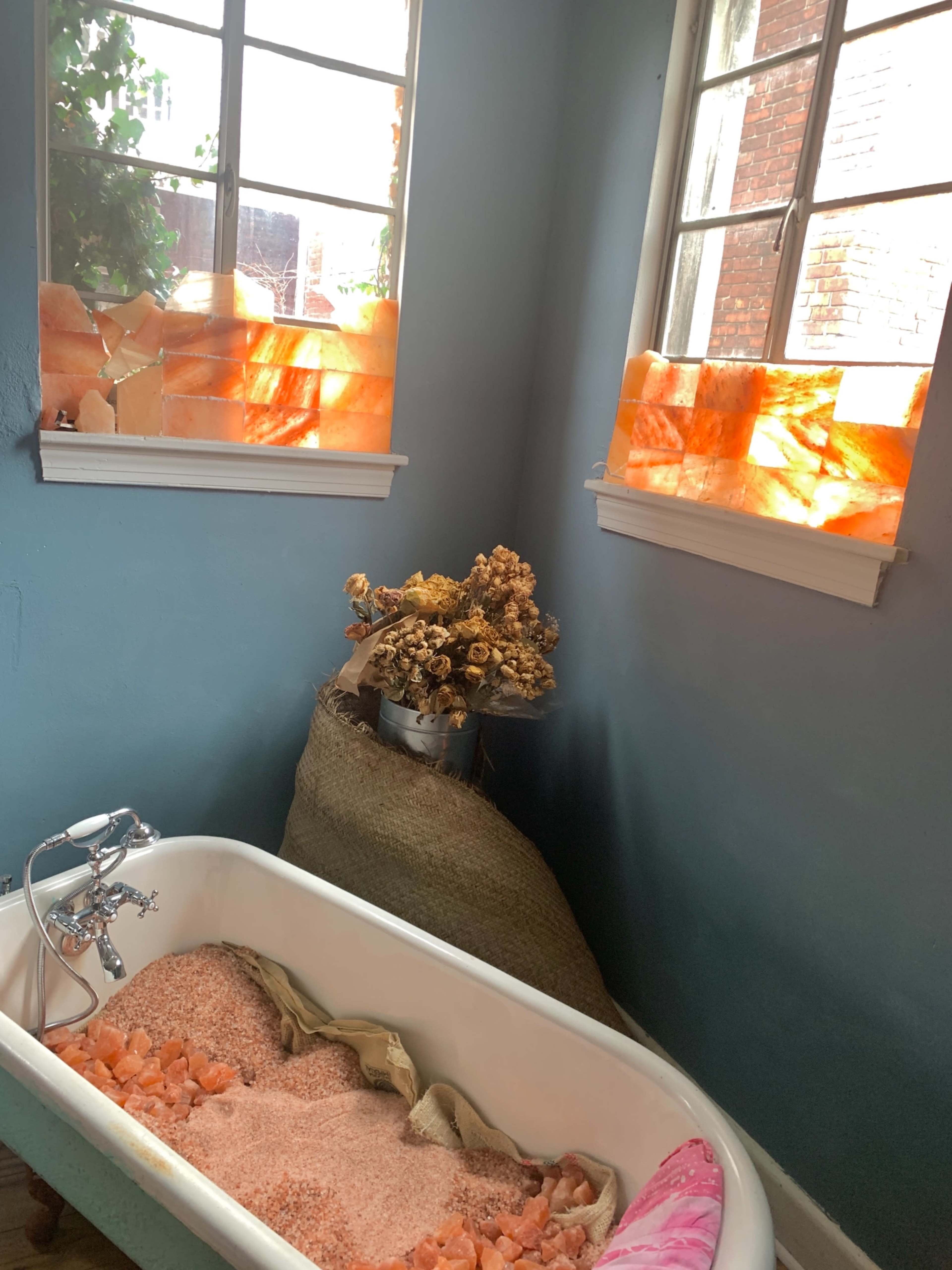 A vintage bathtub filled with pink salt, positioned in a corner of a room with blue walls and large windows adorned with orange-hued materials.