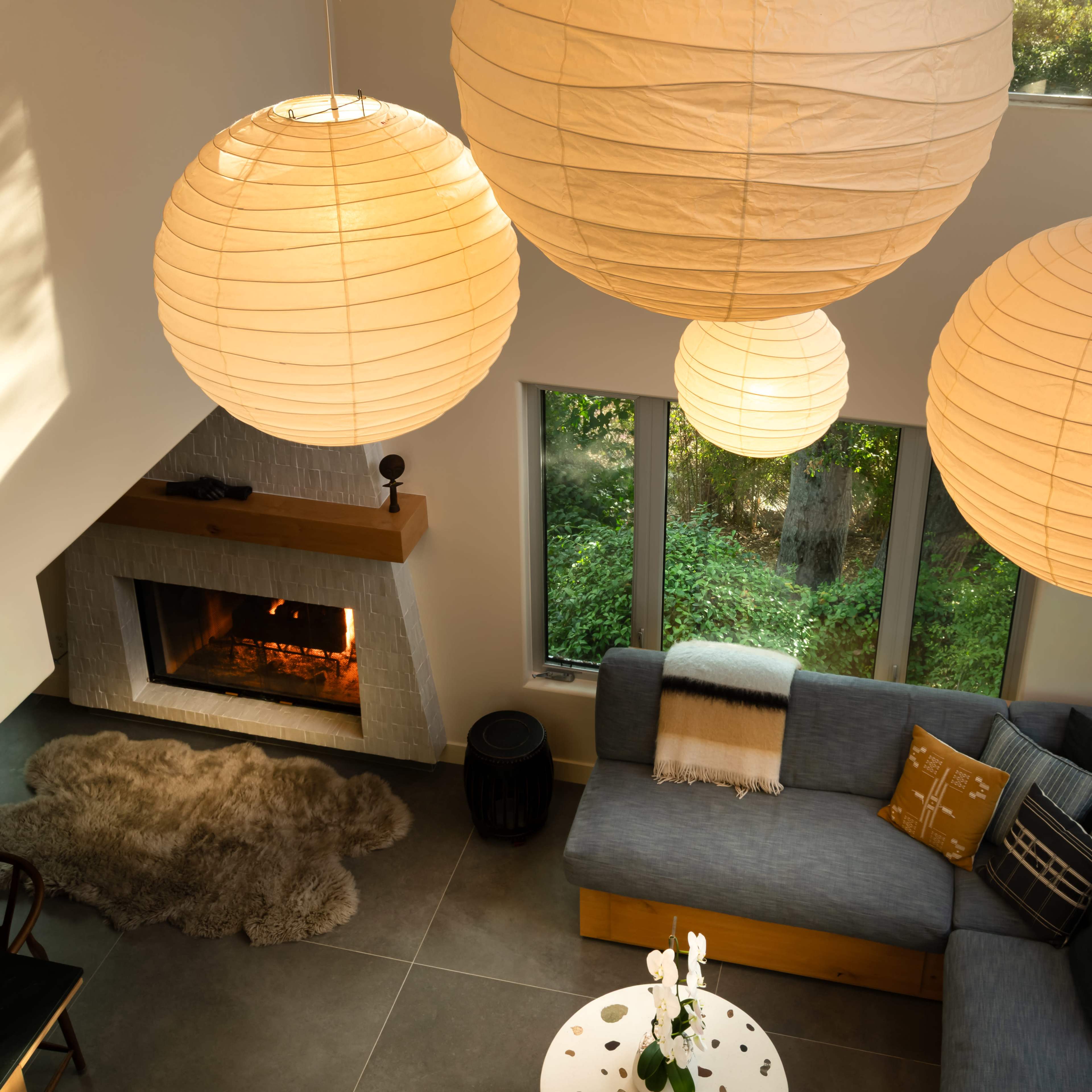 The image shows a modern living room with large paper lanterns hanging from the ceiling, a cozy fireplace, a sectional sofa, and a circular coffee table, all surrounded by greenery visible through the windows.