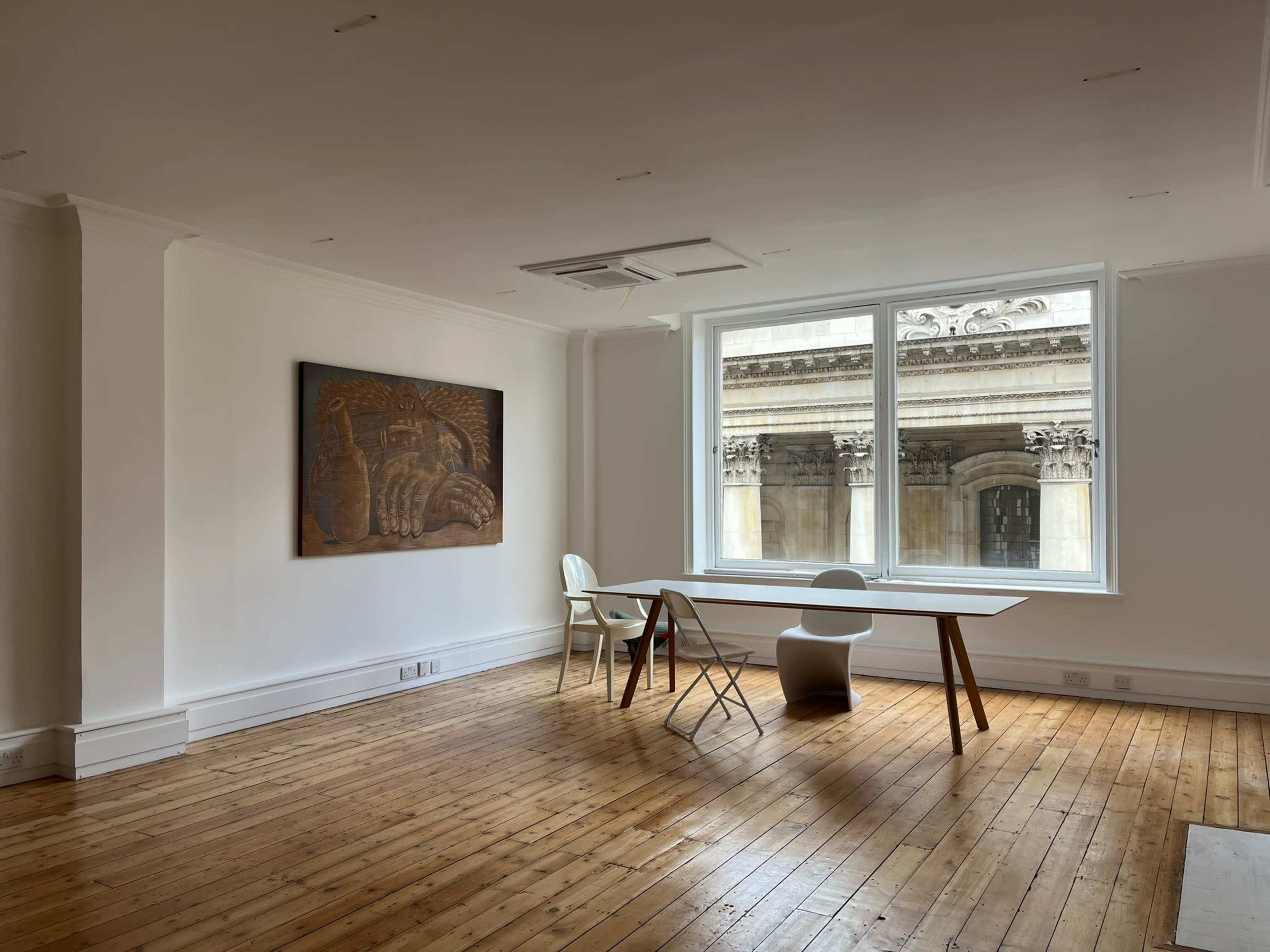 A spacious, empty room with wooden floors, a large window featuring a view of an exterior architectural detail, a wooden table, and a white chair.